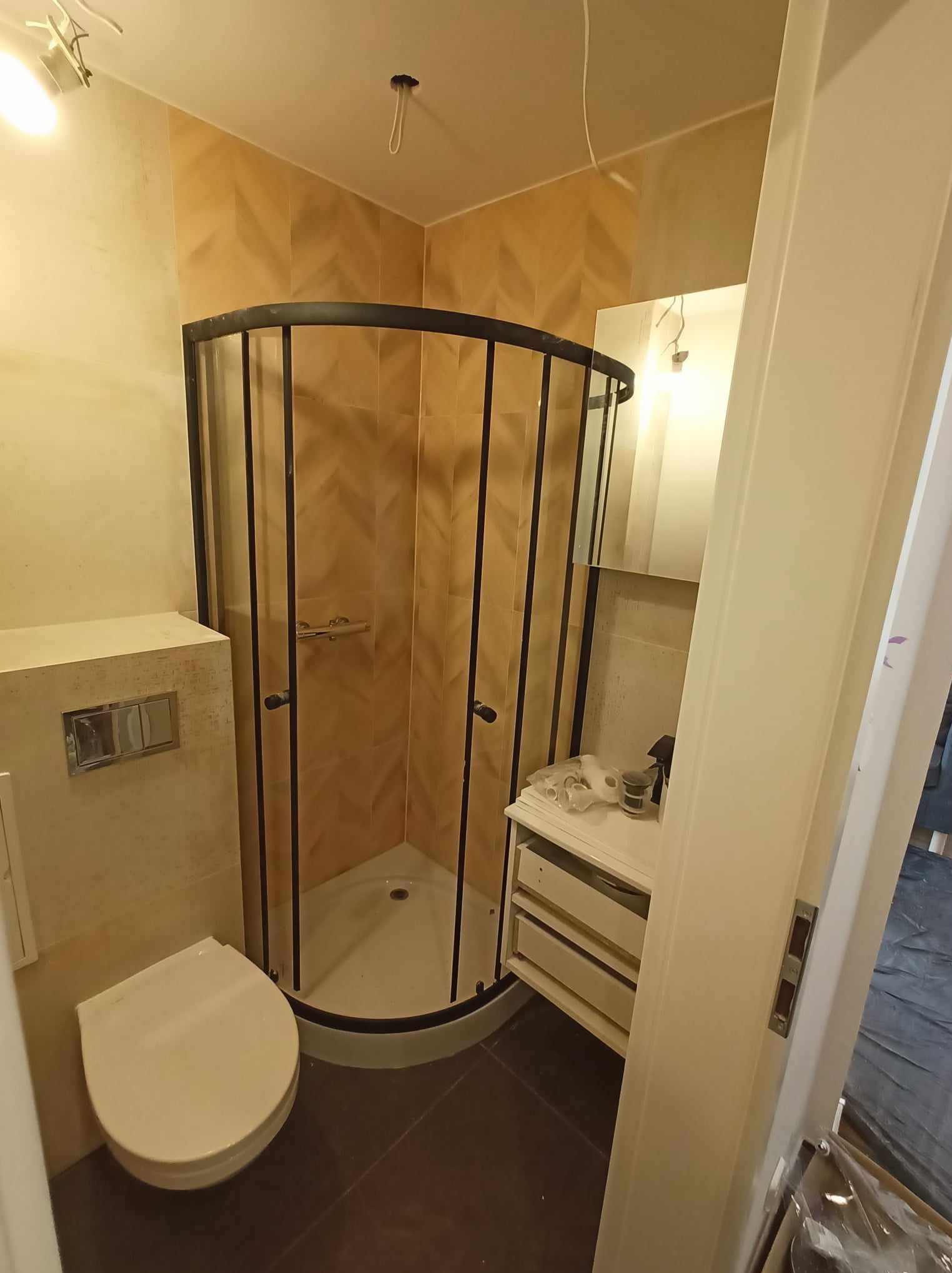 Wykończona łazienka z kabiną prysznicową z czarną ramą, toaletą z podtynkową spłuczką i szafką pod umywalką. Beżowe płytki na ścianach i ciemna podłoga.