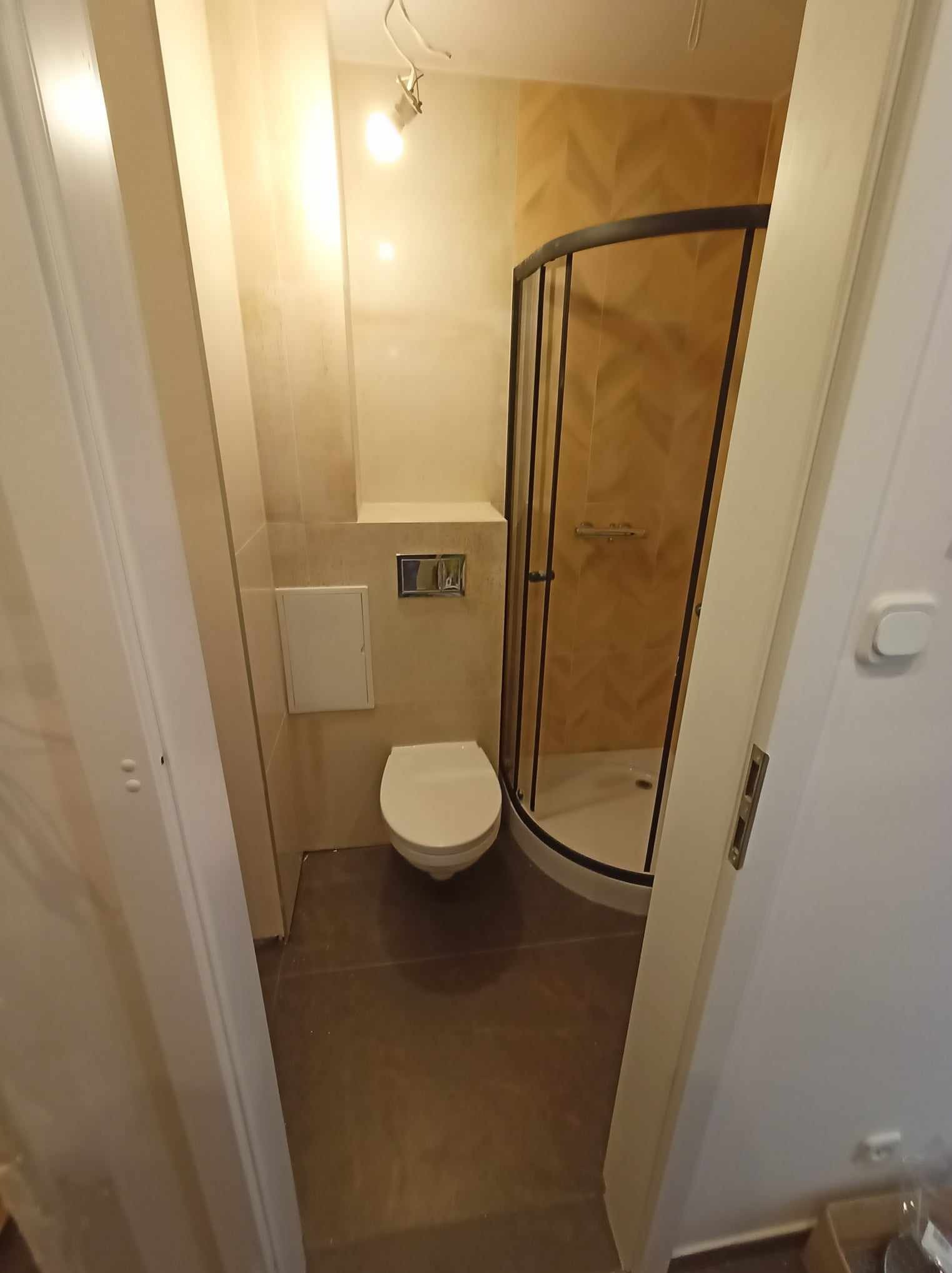 Wykończona łazienka z kabiną prysznicową z czarnymi ramami, toaletą i beżowymi płytkami na ścianach. Widok zza otwartych, białych drzwi.