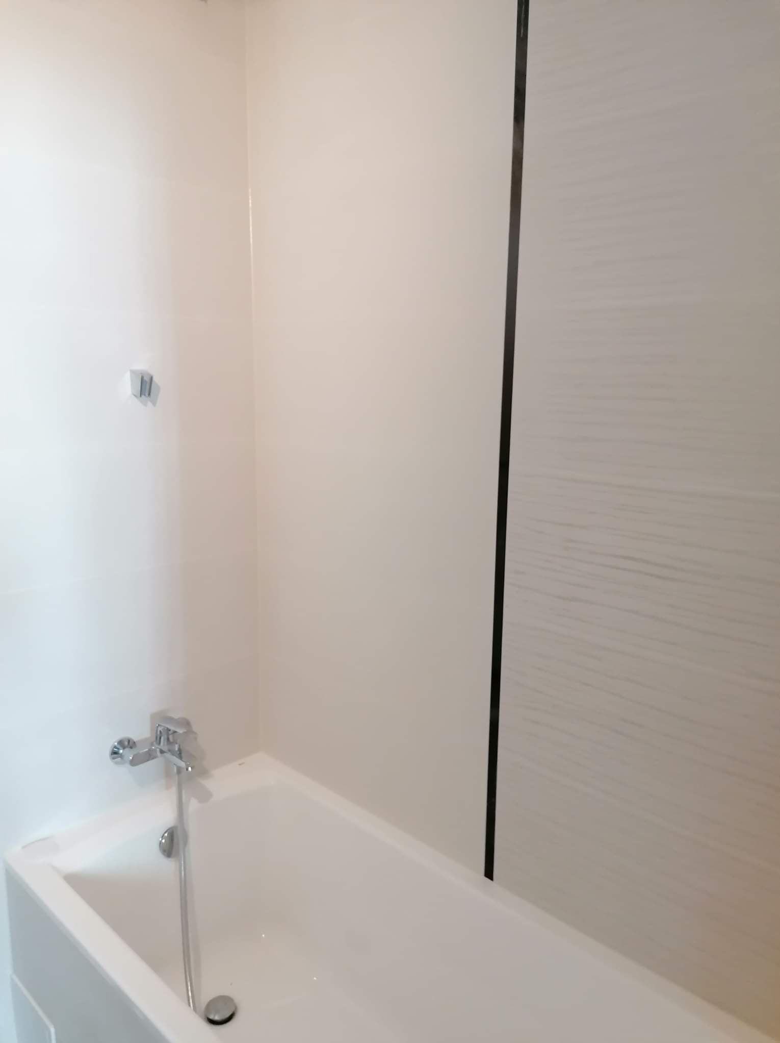 Biała wanna z chromowaną baterią w wykończonej łazience z jasnymi płytkami i panelem ściennym. Minimalistyczny design, czyste linie.