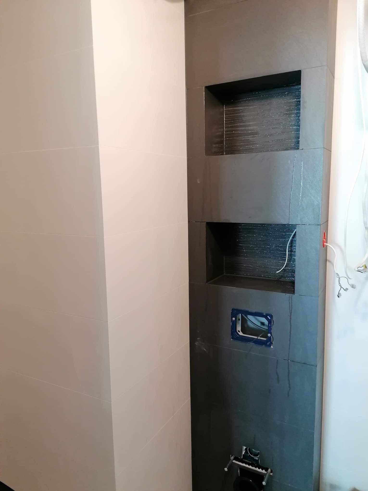 Wykończona wnęka z półkami w łazience, obłożona ciemnymi płytkami. Widoczne przygotowane miejsce na instalację sanitarną i fragment białej ściany z jasnymi płytkami.