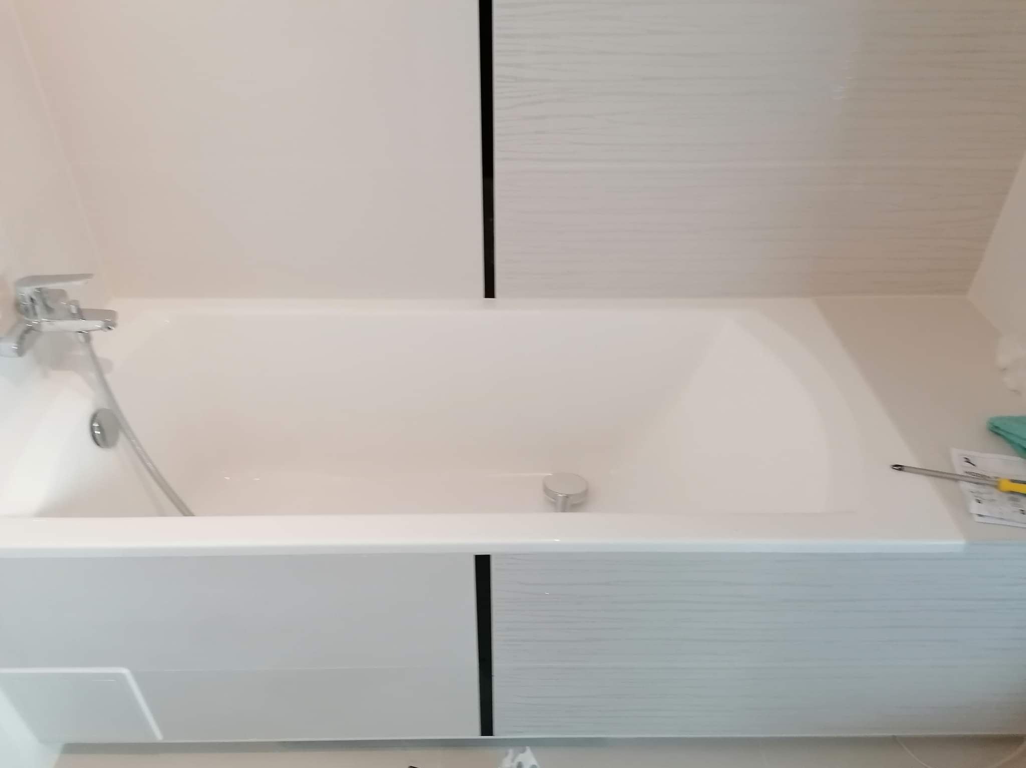 Biała wanna z chromowaną baterią i szarą obudową z czarnymi wstawkami. Na obudowie leżą narzędzia i ściereczka. Widok na wykończoną łazienkę.