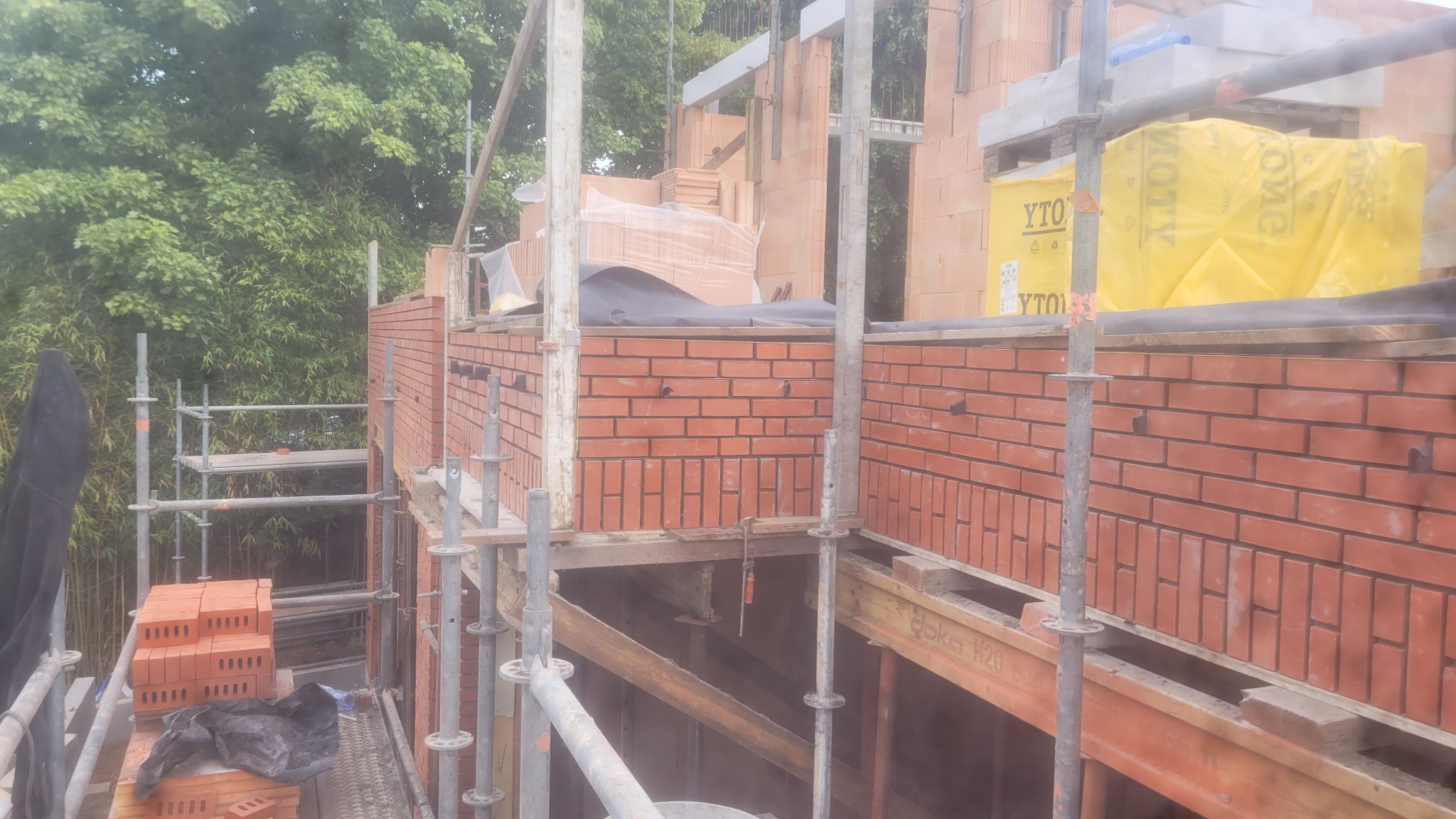 Wznoszenie muru z czerwonej cegły na budowie domu. Widoczne rusztowania, paleta z bloczkami Ytong i fragmenty konstrukcji. Ujęcie z poziomu rusztowania, zieleń w tle.
