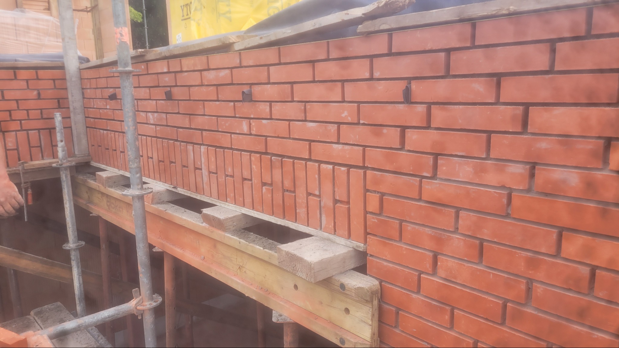 Ściana z czerwonej cegły w trakcie budowy, widoczne rusztowania i podpory. Cegły ułożone w regularny wzór, z widocznymi spoinami. Częściowo widoczna dłoń.