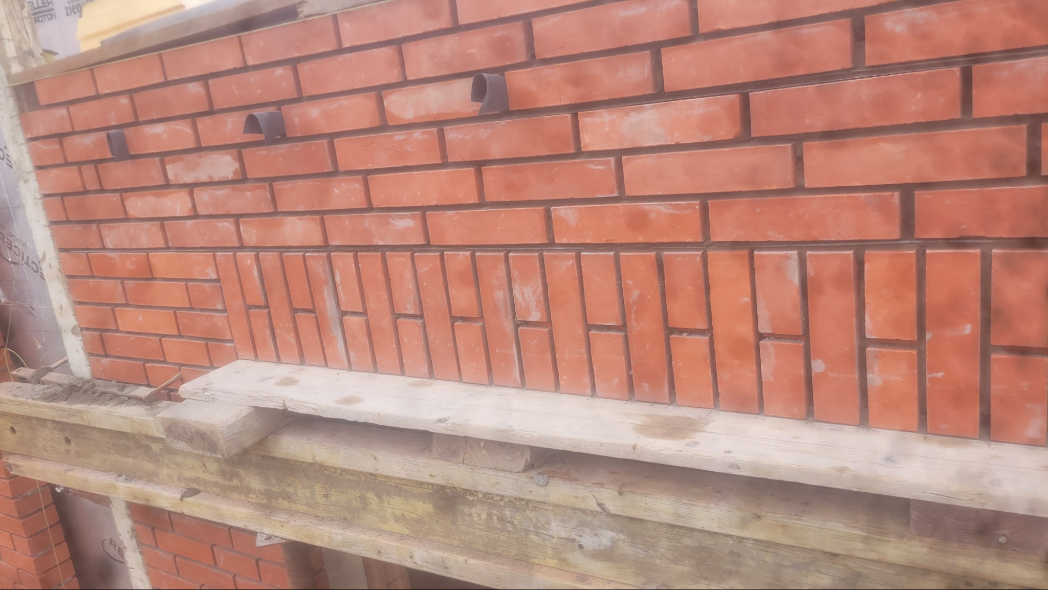 Ceglana ściana z dekoracyjnym układem cegieł w pionie, wsparta drewnianą konstrukcją. Widoczne fragmenty instalacji i materiałów budowlanych.