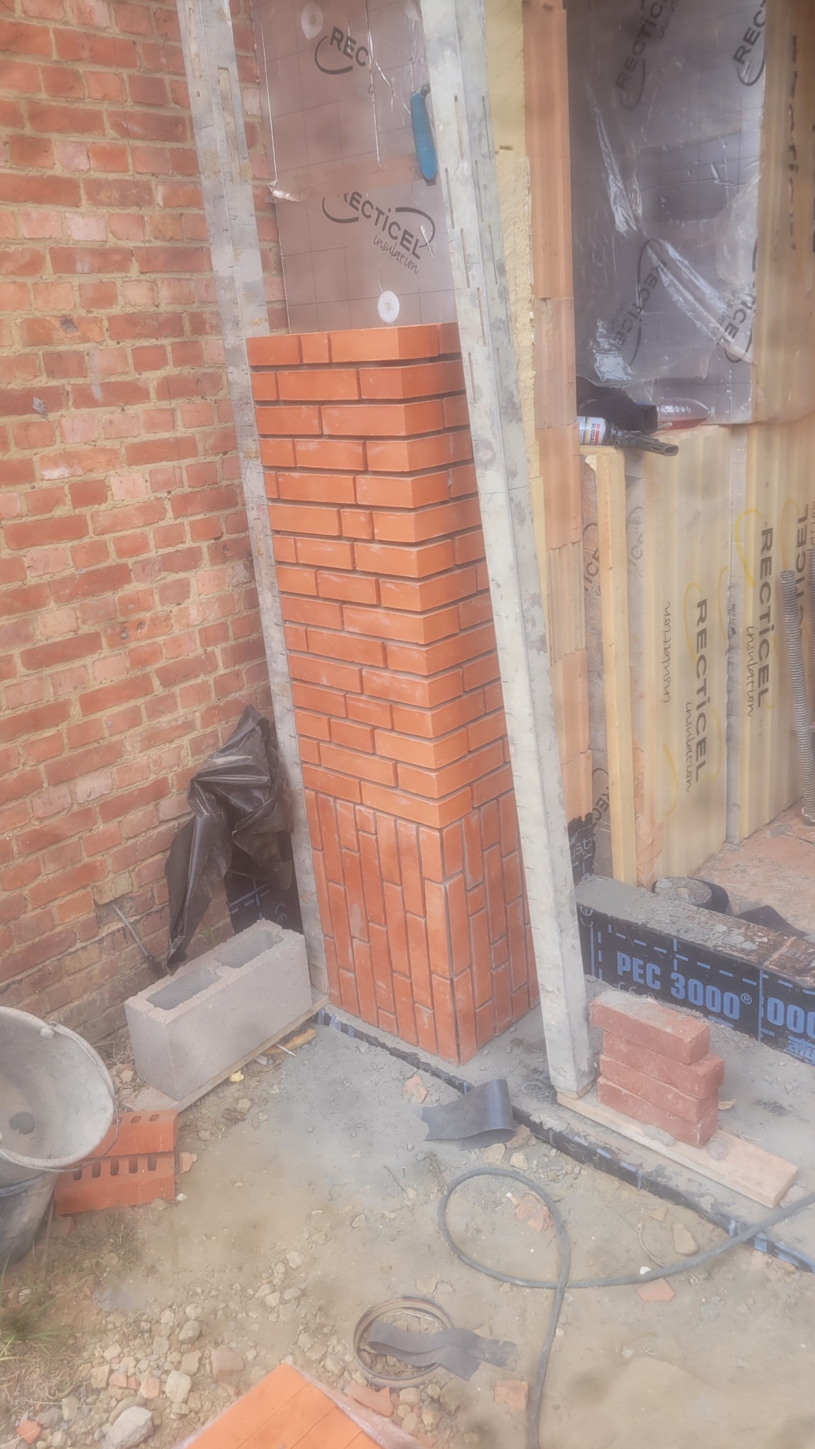 Wznoszenie muru z czerwonej cegły, widoczne fragmenty ściany z cegły, izolacji i narzędzi budowlanych. Prace murarskie w trakcie realizacji.