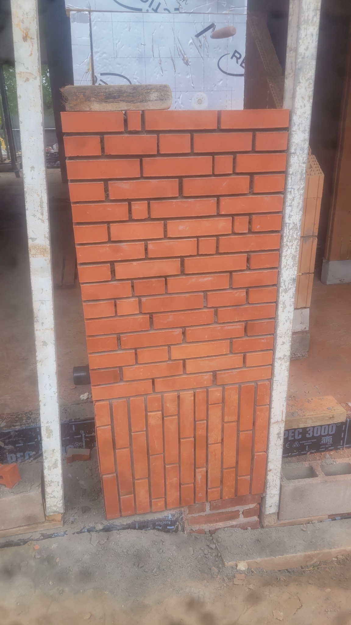 Fragment nowej ściany z czerwonej cegły, z widocznym wzorem ułożenia cegieł. Białe, surowe ramy okienne po bokach. Widoczne materiały budowlane w tle.