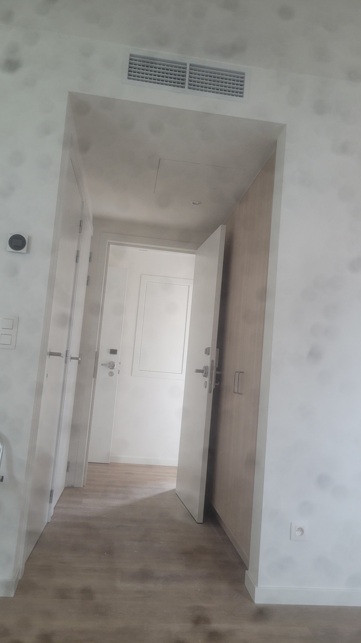 Jasne wnętrze z otwartymi drzwiami i widoczną wentylacją sufitową. Minimalistyczny design, świeżo po remoncie, z drewnopodobną podłogą i białymi ścianami.