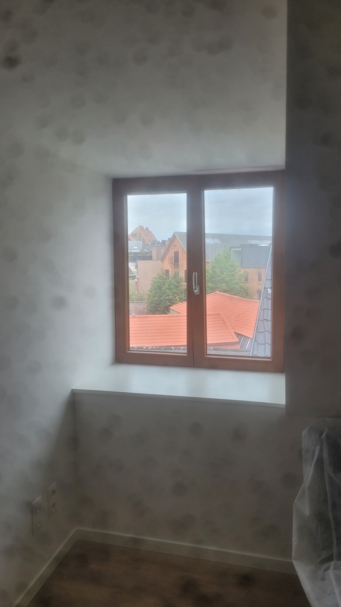 Pokój po odświeżeniu: drewniane okno z widokiem na dachy budynków, świeżo pomalowane ściany w odcieniach bieli, widoczne gniazdka elektryczne i fragment podłogi.