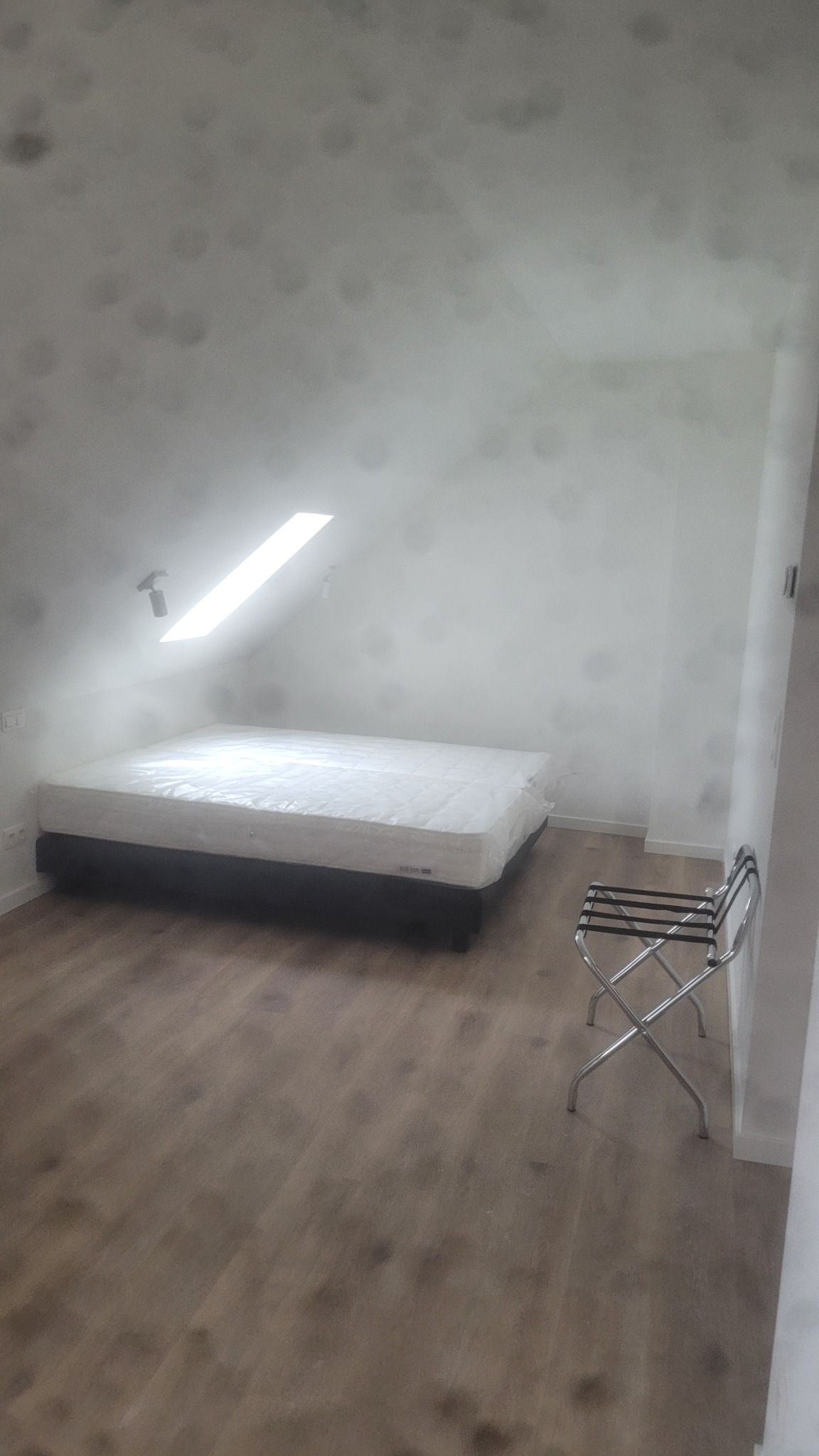 Pokój na poddaszu z łóżkiem i drewnianą podłogą. Ściany białe, widoczne ślady wilgoci. Metalowy stojak na walizki przy ścianie. Okno dachowe wpuszcza jasne światło.