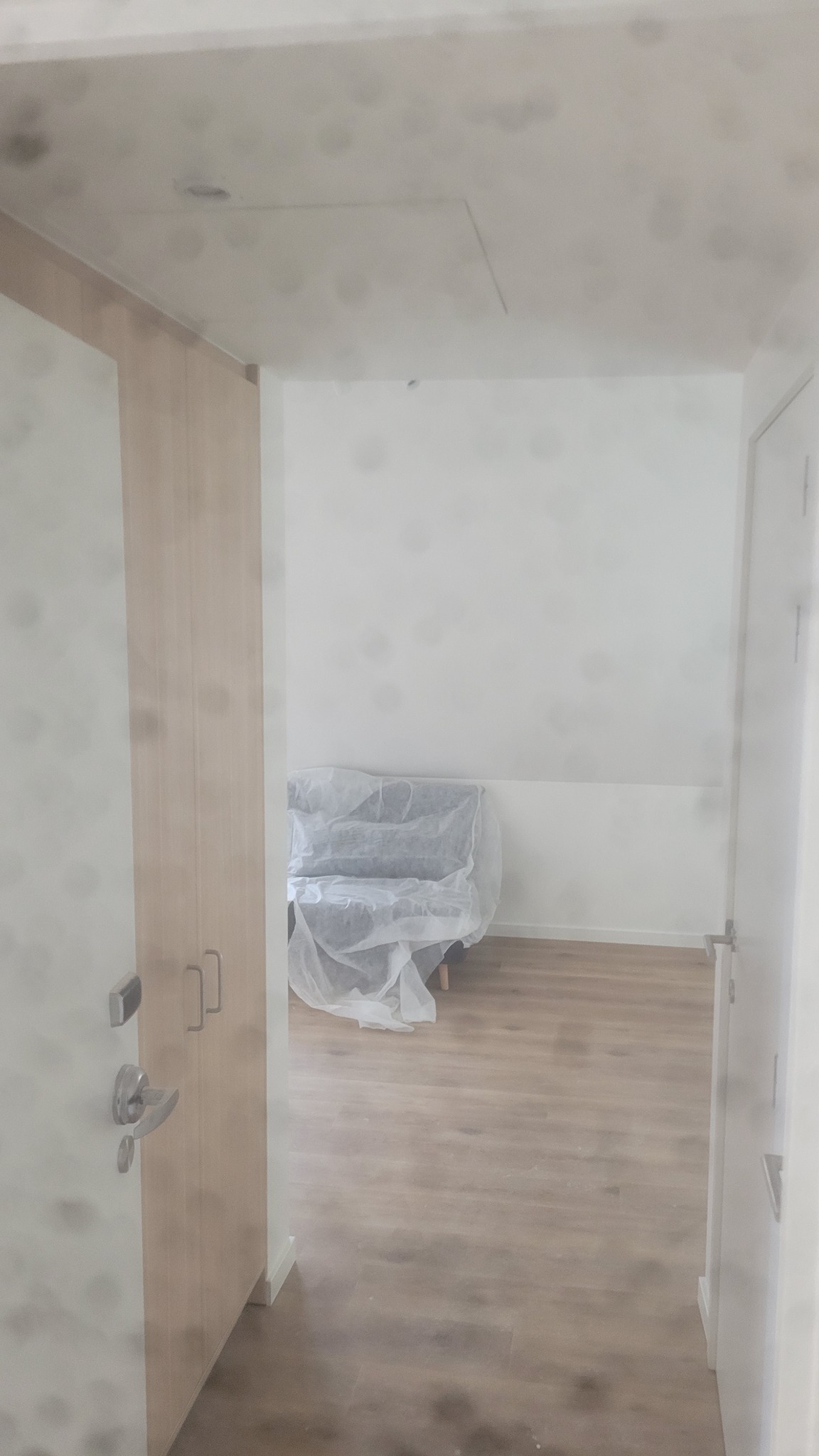 Jasne wnętrze po remoncie: parkiet, białe ściany, meble zabezpieczone folią. Widok z korytarza, drzwi po prawej, szafa po lewej. Minimalistyczny styl.