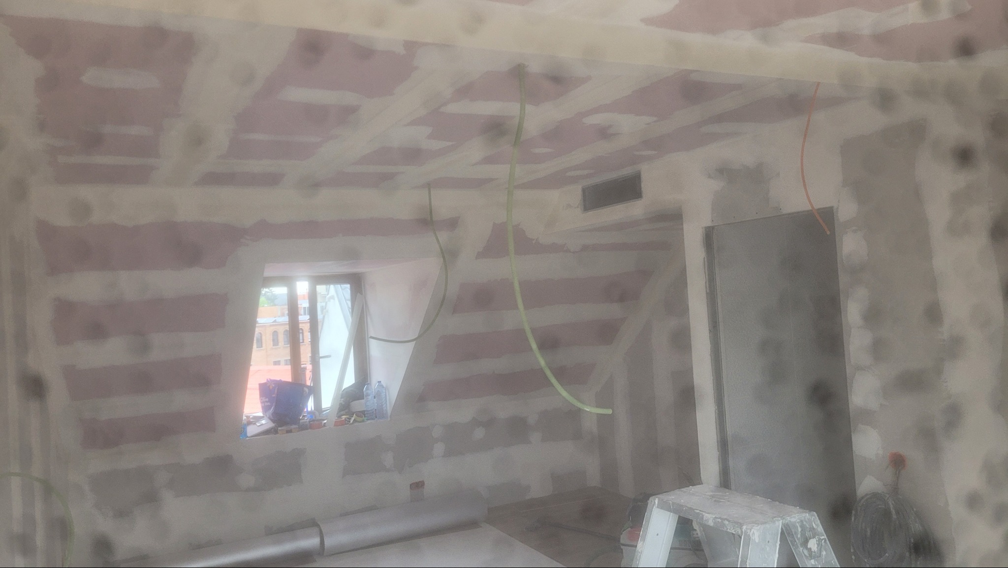 Poddasze w trakcie remontu: widoczne płyty karton-gips na skosach, szpachlowane łączenia, wystające kable i drabina malarska. Okno z widokiem na budynki.
