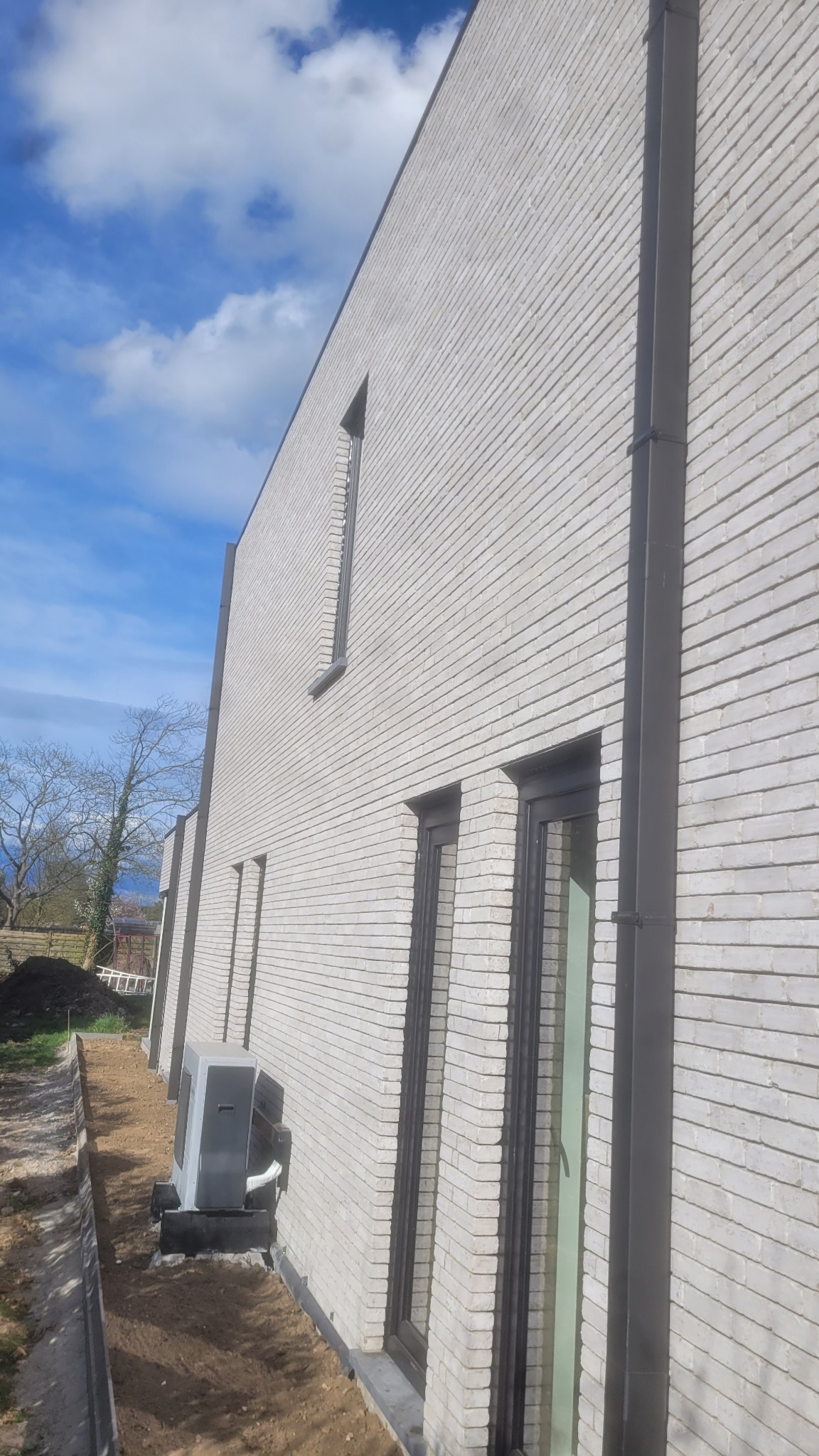 Elewacja budynku z jasnej cegły z ciemnymi oknami i rurą spustową. Widoczny klimatyzator przy ścianie i fragment nieba z chmurami. Ujęcie z dołu, podkreślające wysokość budynku.