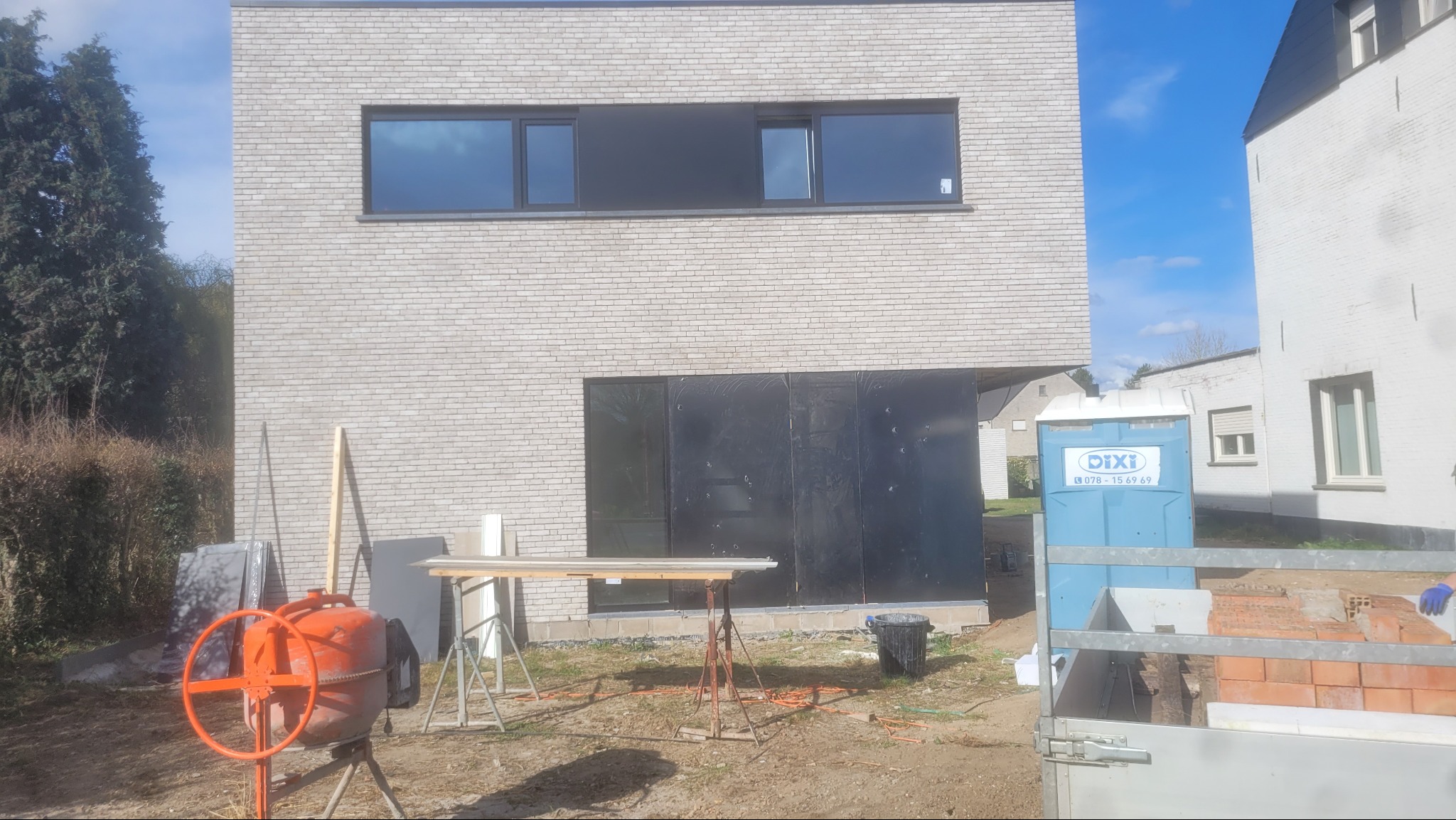 Nowoczesny dom w trakcie budowy: jasna cegła, czarne okna, zabezpieczone otwory. Na pierwszym planie betoniarka i materiały budowlane, przenośna toaleta w tle.