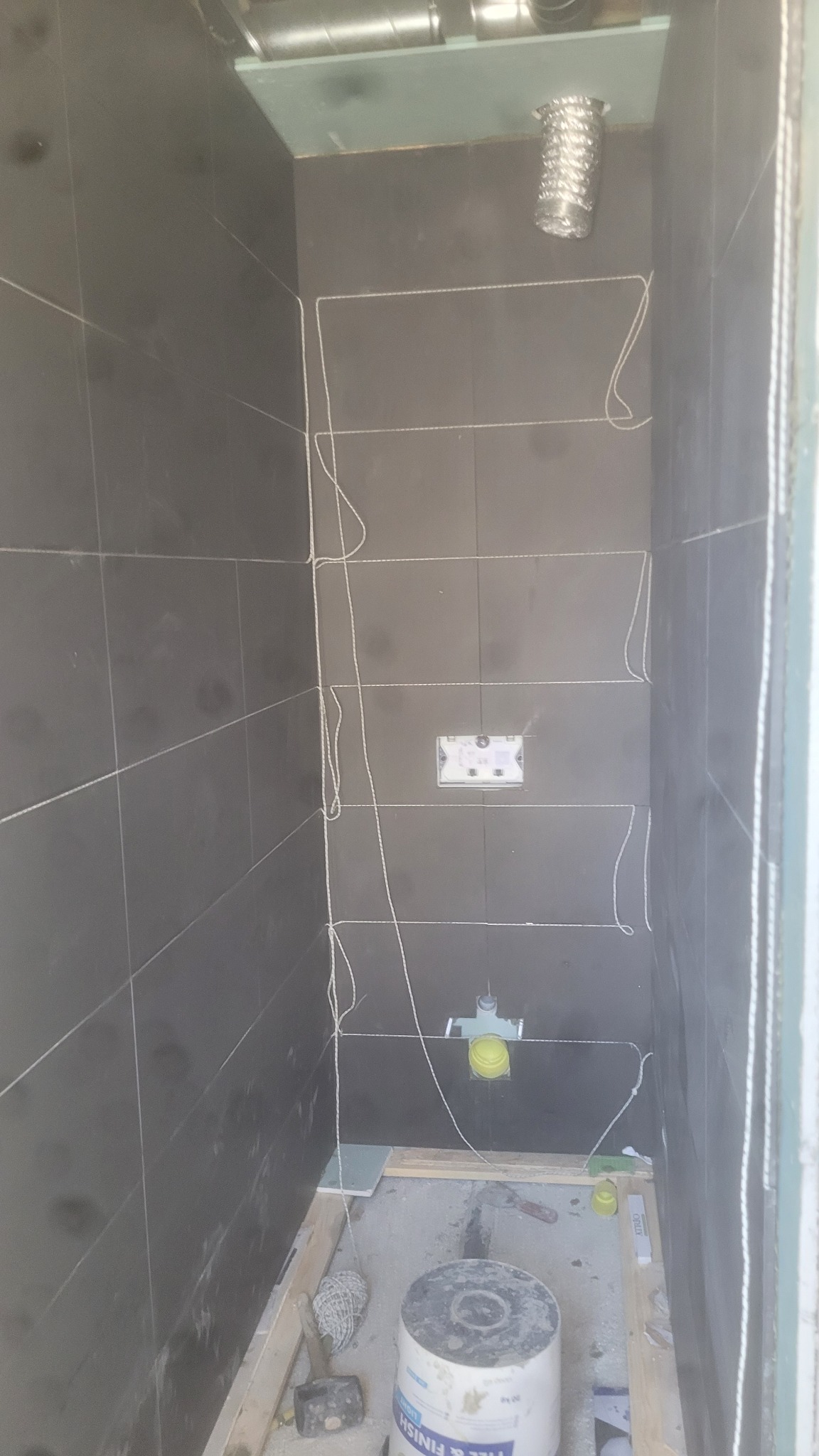 Ciemne płytki ścienne w trakcie montażu w małej łazience. Widoczne przewody, puszka elektryczna, rury i narzędzia glazurnicze. W tle wentylacja.