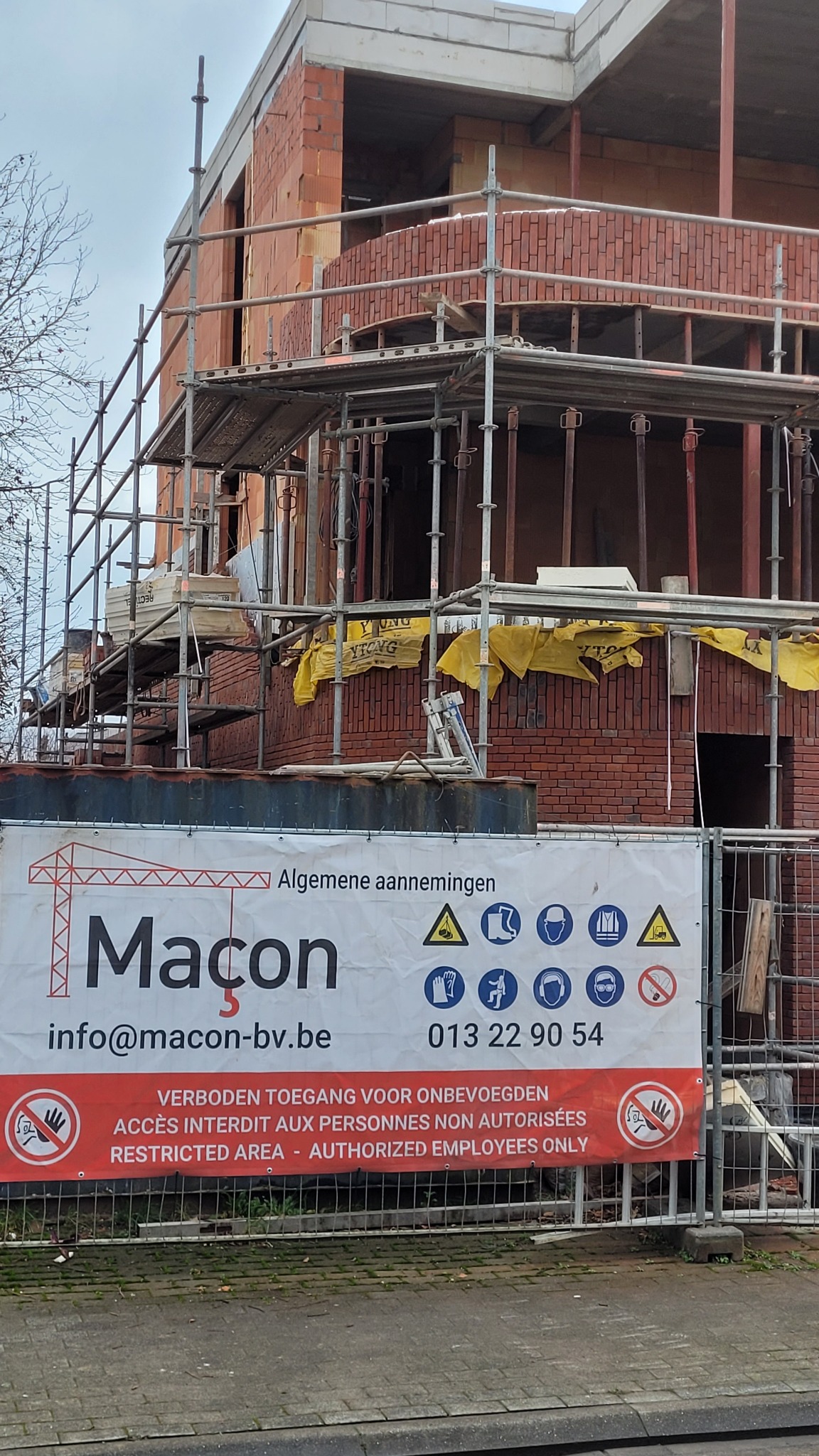 Budowa domu z czerwonej cegły w trakcie realizacji, widoczne rusztowania i baner informacyjny firmy budowlanej z nazwą 'Macon' i danymi kontaktowymi.