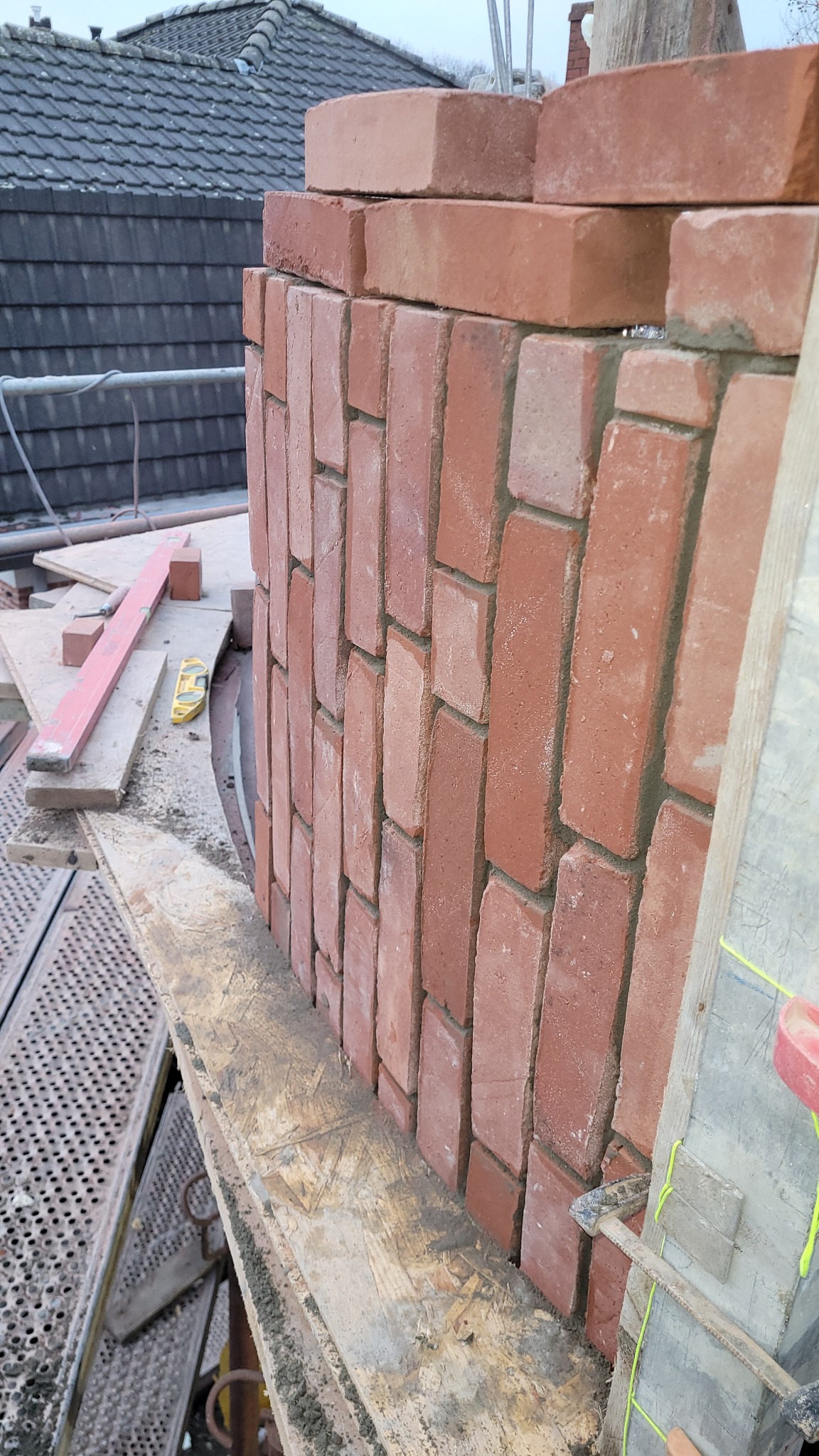 Ceglana ściana w trakcie murowania, widoczna z rusztowania. Czerwona cegła ułożona w regularny wzór, z widocznymi spoinami. W tle dach budynku i narzędzia murarskie.