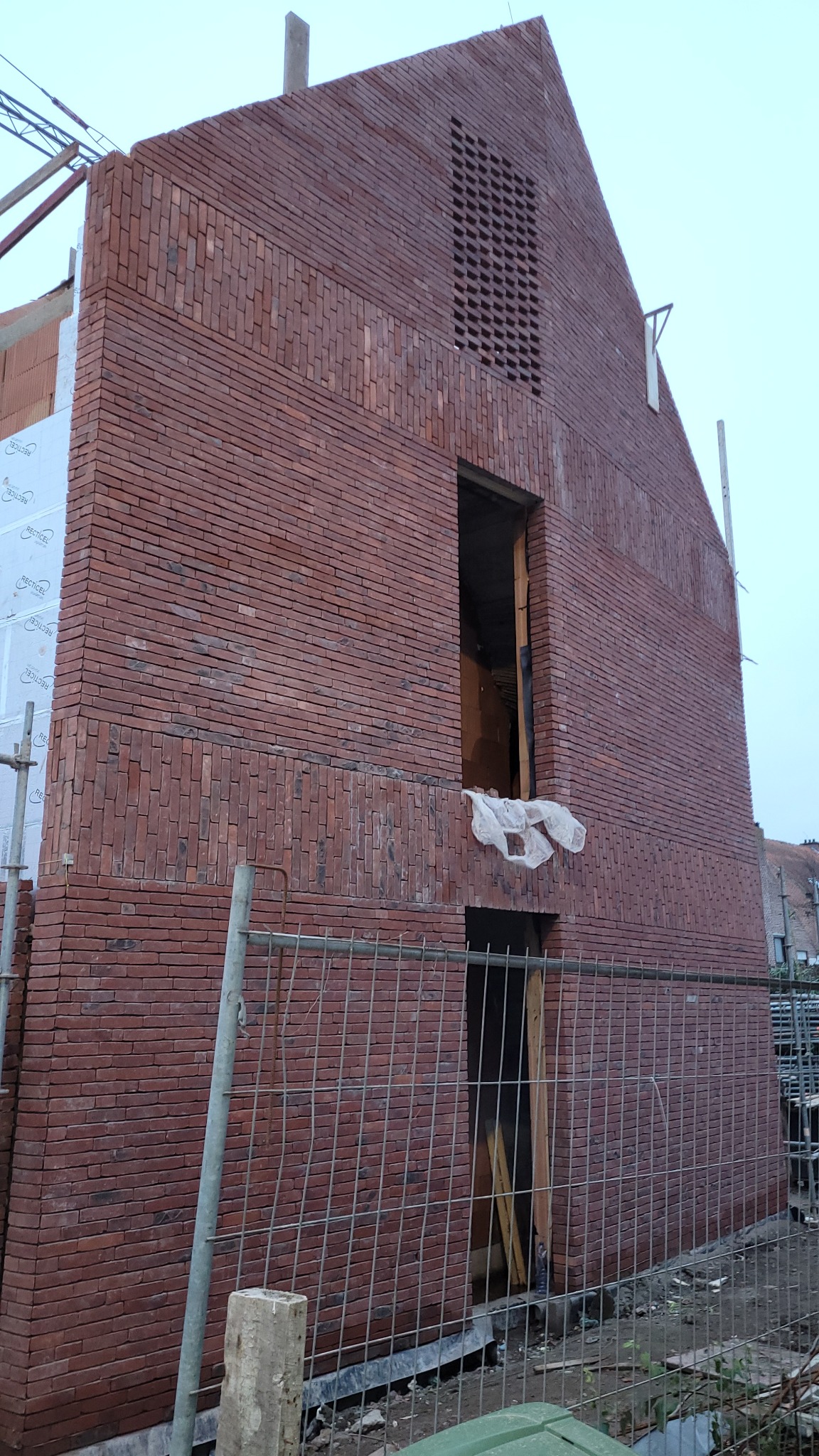 Wznoszona ściana z czerwonej cegły klinkierowej z ażurowym fragmentem i otworami okiennymi, zabezpieczona siatką budowlaną. Widoczny fragment ocieplenia.