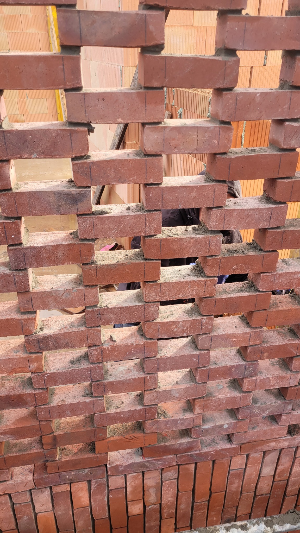 Ażurowa ściana z czerwonej cegły, widok z bliska, z widocznymi spoinami i fragmentem osoby w tle. Nietypowy układ cegieł tworzy dekoracyjny wzór.