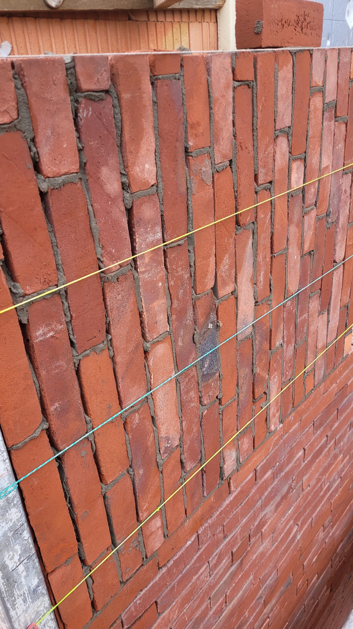 Ceglana ściana w trakcie murowania, z widocznymi sznurami poziomującymi, Śrem. Czerwona cegła i szara zaprawa tworzą surowy, budowlany krajobraz.