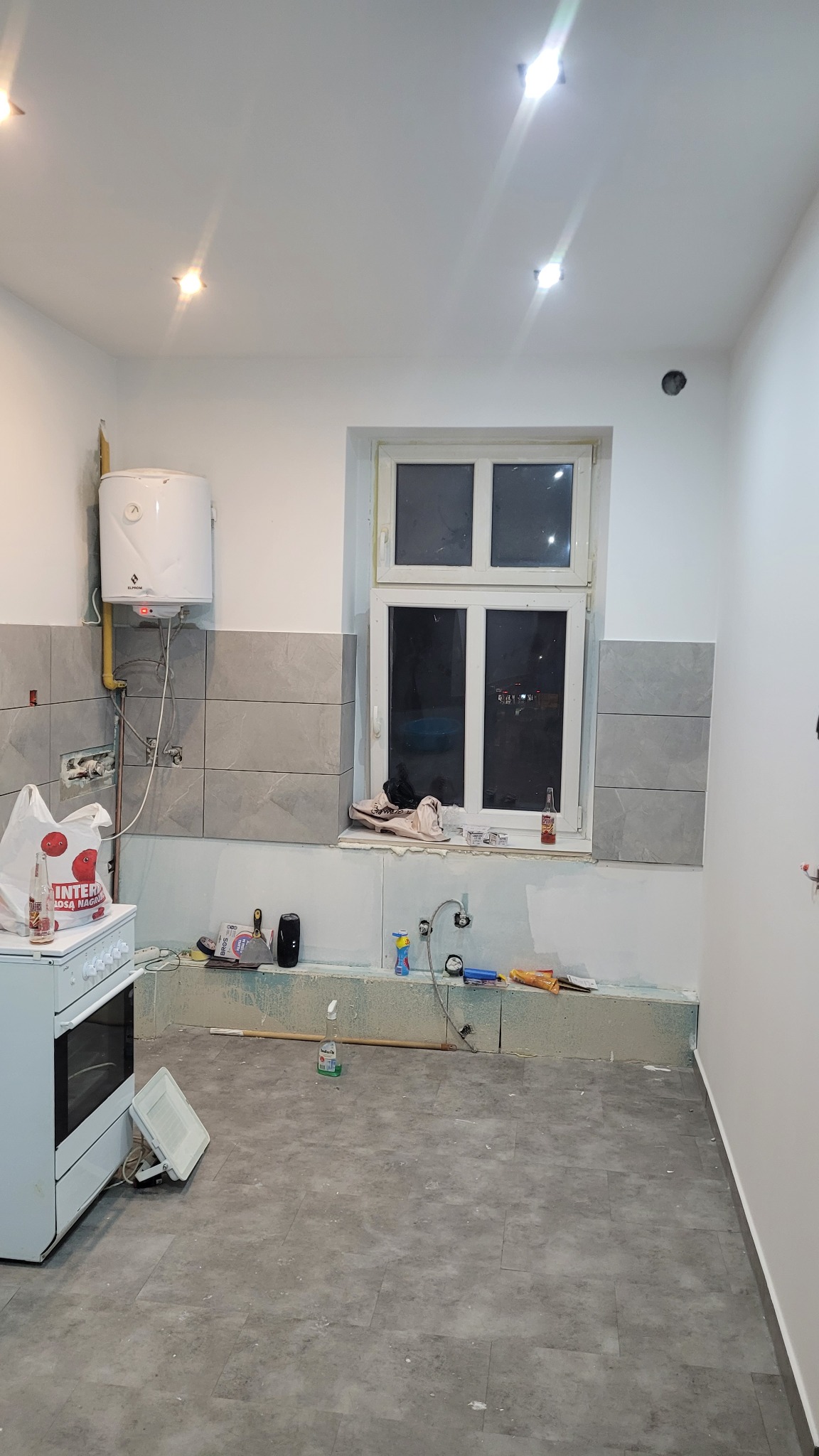 Kuchnia w trakcie remontu: szare płytki na ścianie, biały bojler, okno z widokiem na noc, częściowo położona podłoga z szarych płytek. Widoczne narzędzia i akcesoria.