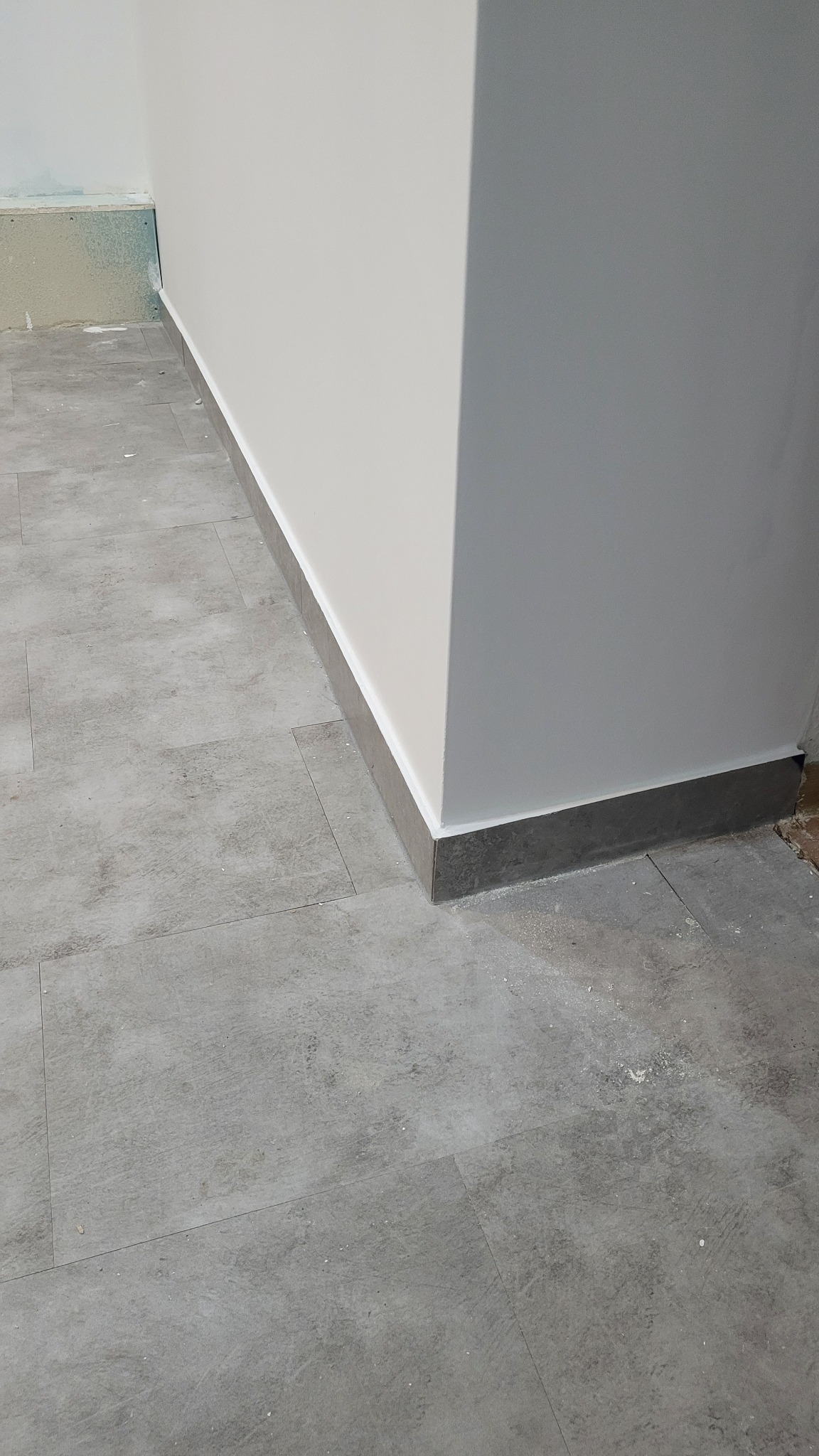 Szare płytki podłogowe imitujące kamień, ułożone przy ścianie pomalowanej na biało z szarą listwą przypodłogową. Widoczny fragment ściany z niedokończonym wykończeniem.