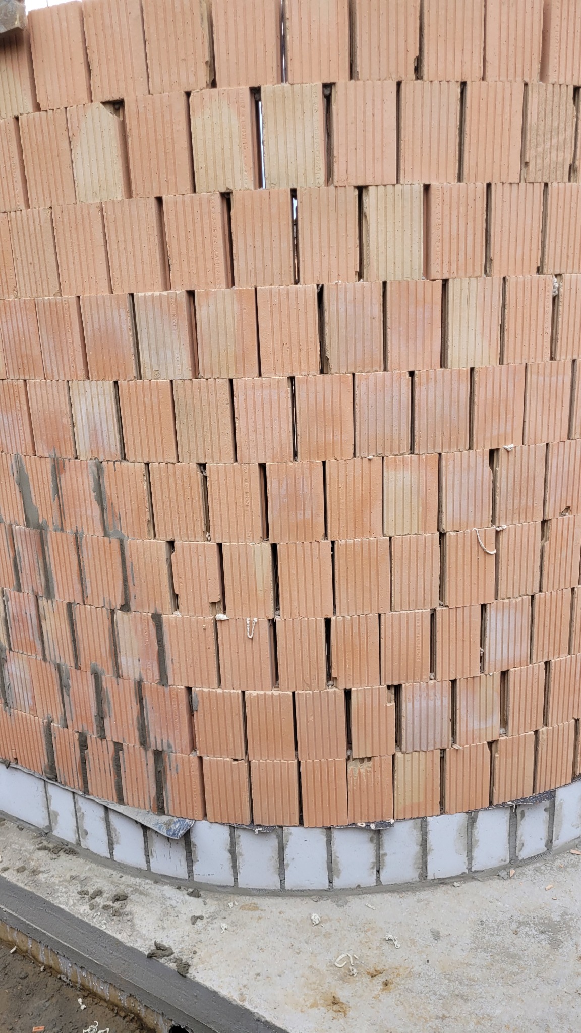 Wznoszenie okrągłej ściany z czerwonych, porowatych pustaków ceramicznych na fundamencie z białych bloczków. Widoczne spoiny i struktura materiałów.