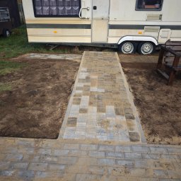 SebBruk - Ścieżka z kostki brukowej prowadząca do przyczepy kempingowej, z widocznymi wykopami po bokach i piaskiem na kostce, sugerująca świeżo zakończone prace brukarskie.