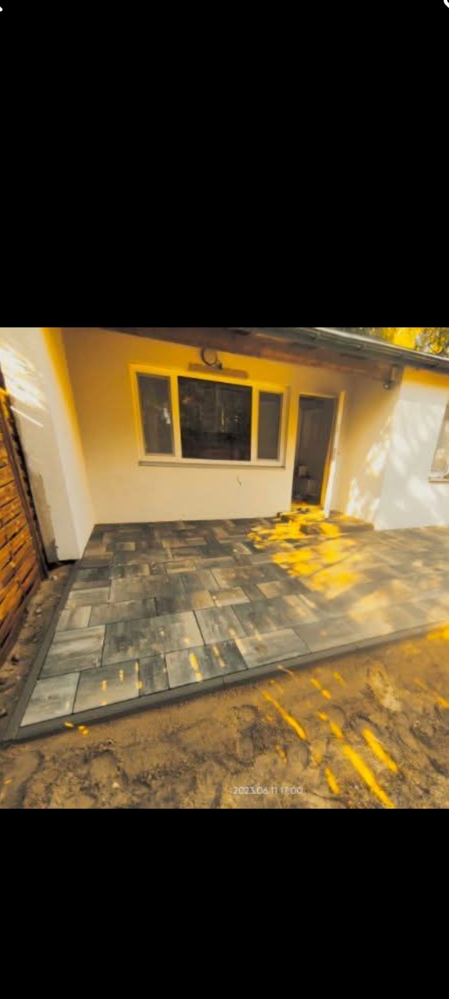 Ułożona kostka brukowa w odcieniach szarości tworzy taras przy budynku z białymi ścianami. Widoczne promienie słoneczne i fragment drewnianego ogrodzenia.