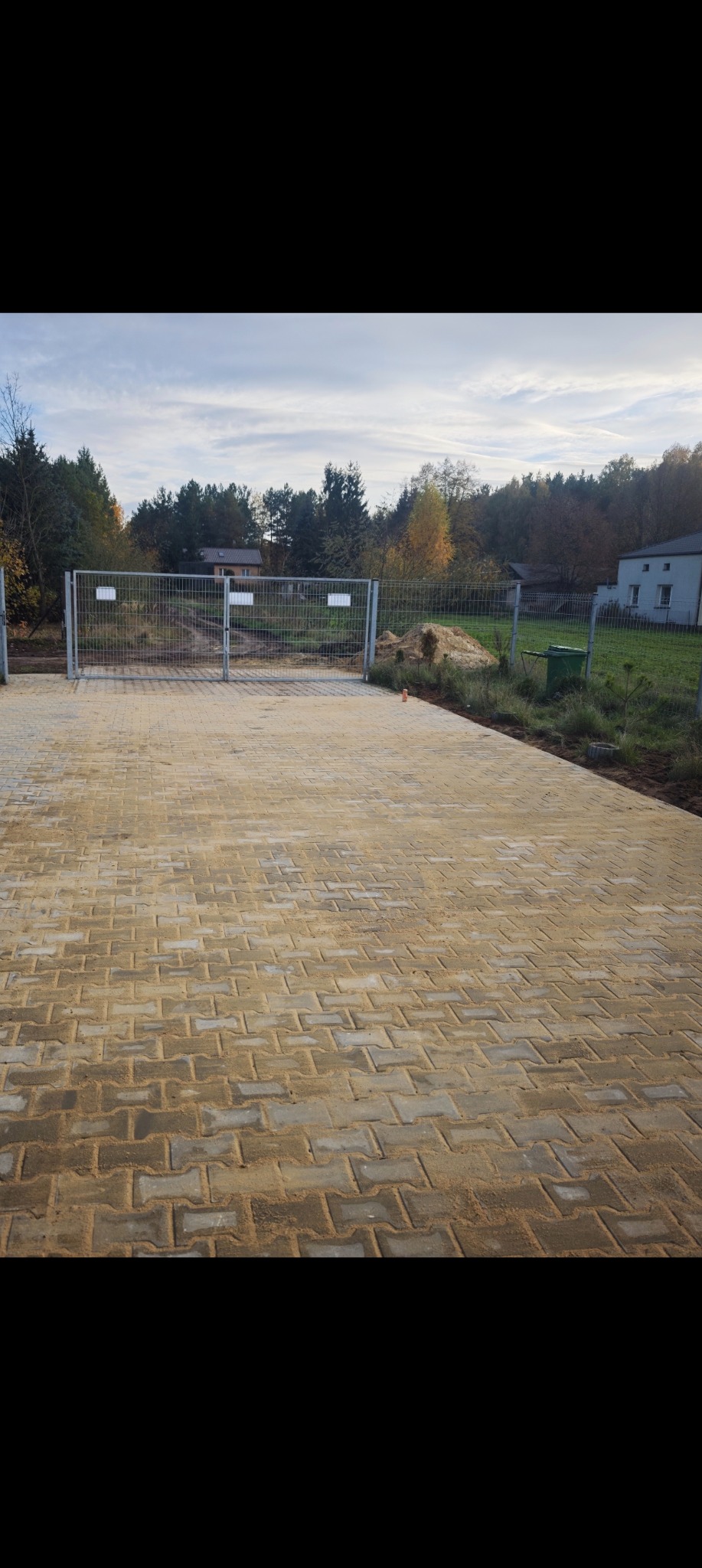 Nowa, beżowa kostka brukowa ułożona na posesji, widok z góry. W tle metalowa siatka ogrodzeniowa i zieleń drzew. Świeżo wykonana nawierzchnia.