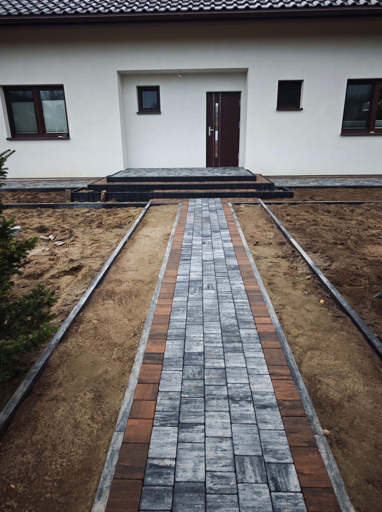 Ścieżka z kostki brukowej prowadząca do domu z brązowymi oknami i drzwiami. Kostka w odcieniach szarości i brązu, obrzeża z szarego betonu. Częściowo wykonana praca.