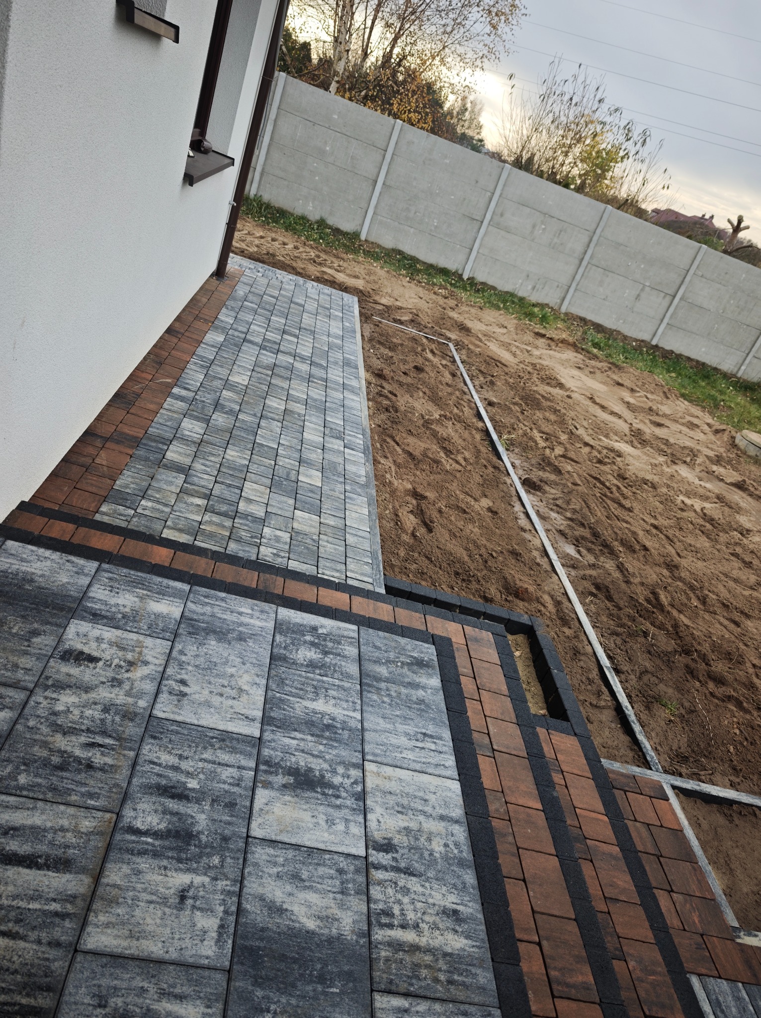 Ułożona kostka brukowa w odcieniach szarości i brązu, obrzeża z ciemnej kostki. Widoczny fragment budynku i ogrodzenie betonowe w tle. Teren przygotowany pod dalsze prace.