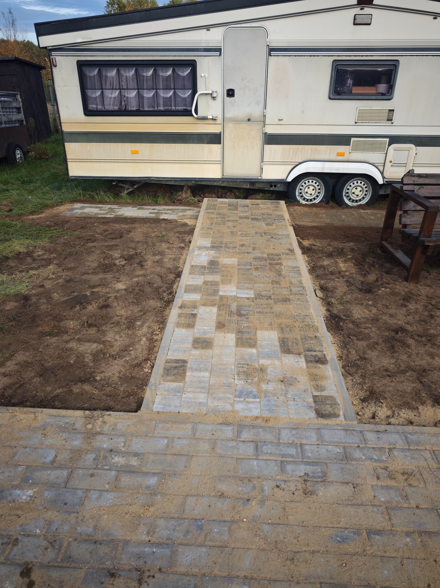 Ścieżka z kostki brukowej prowadząca do przyczepy kempingowej, z widocznymi wykopami po bokach i piaskiem na kostce, sugerująca świeżo zakończone prace brukarskie.
