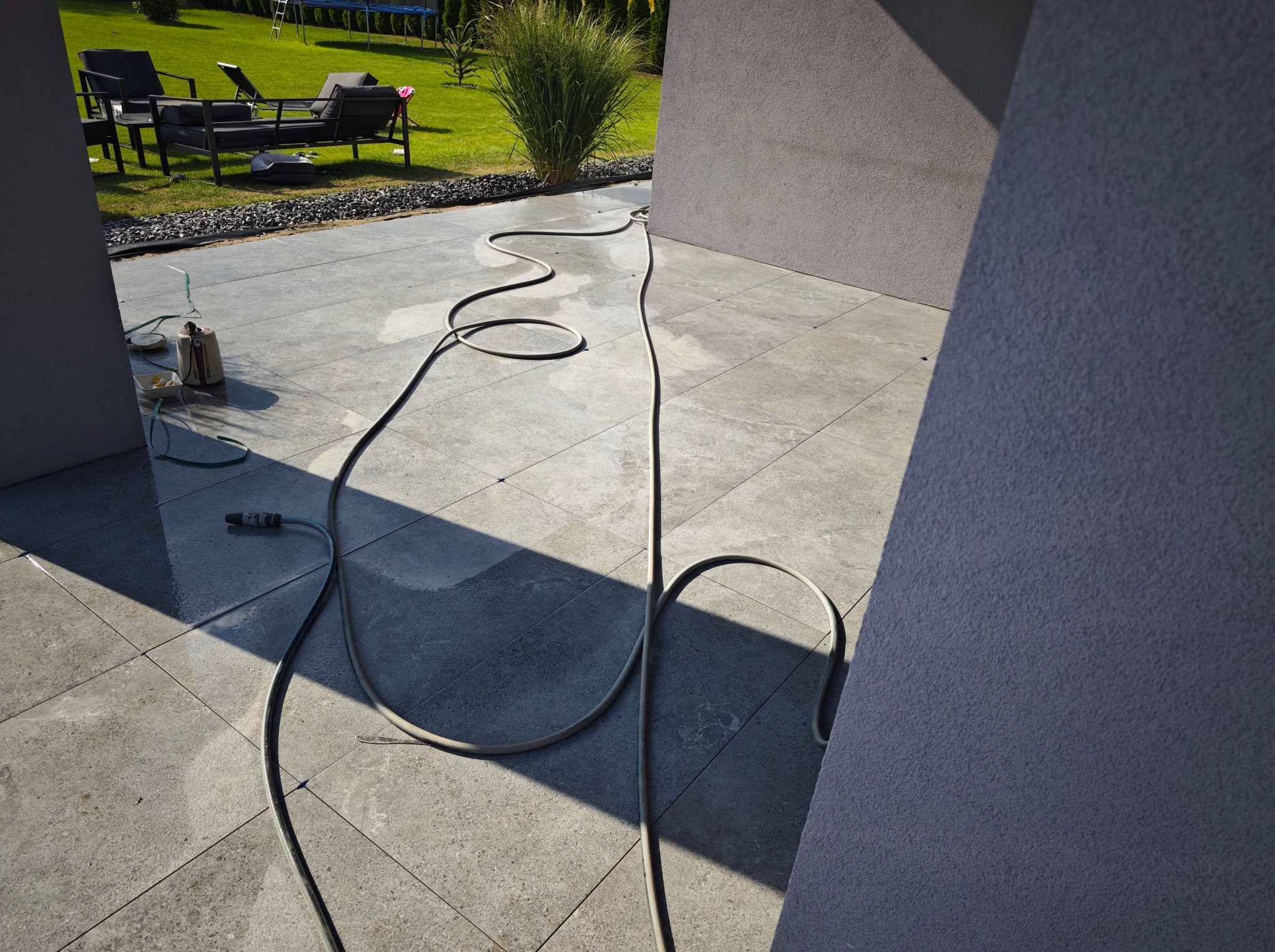 Szara kostka brukowa na tarasie w słoneczny dzień, z widocznym wężem ogrodowym i elementami wyposażenia ogrodu w tle. Widoczne ślady po czyszczeniu kostki.