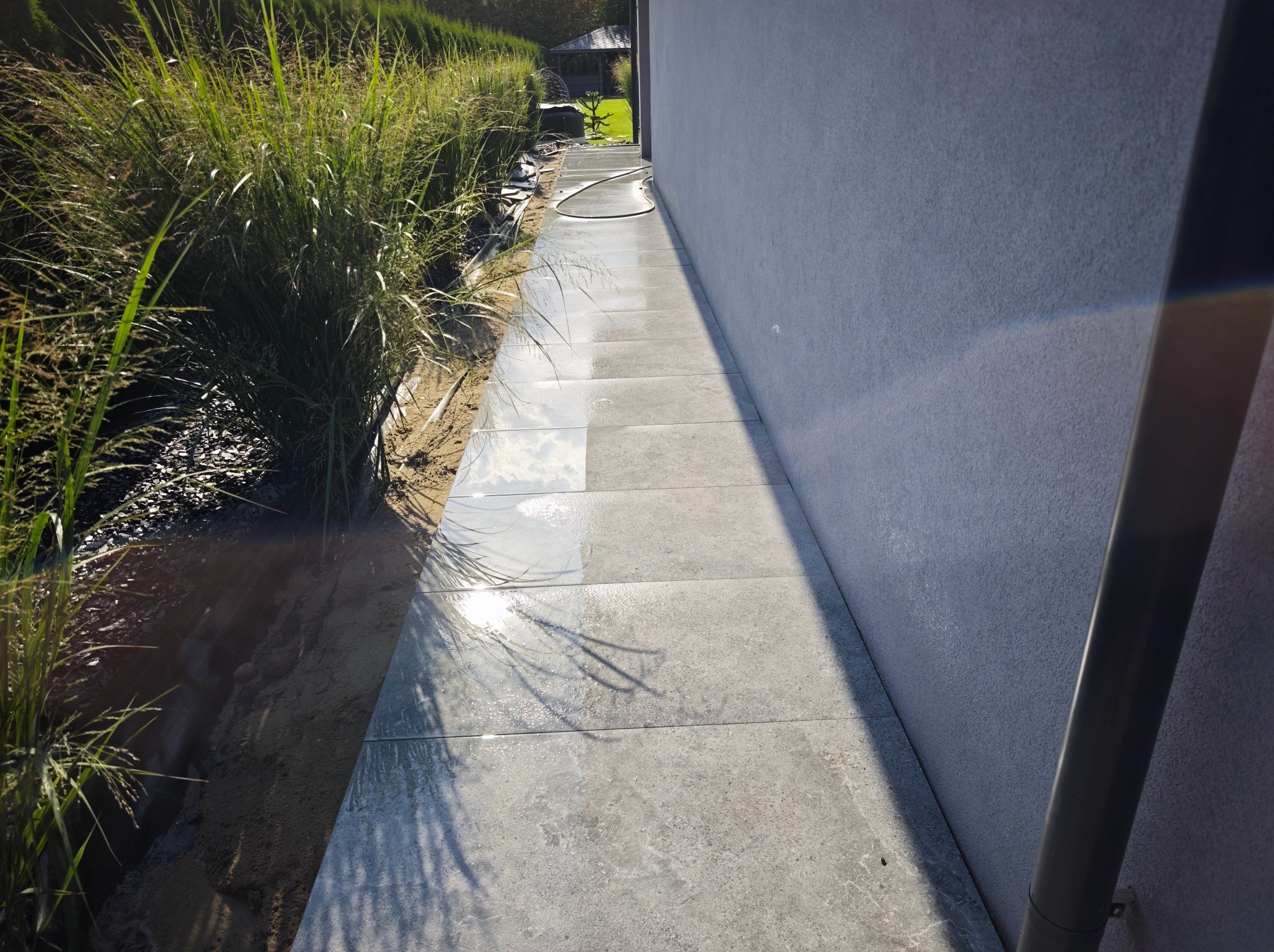 Świeżo ułożony chodnik z szarych płyt betonowych przylegający do ściany budynku, z widoczną roślinnością po lewej stronie i śladami wody po czyszczeniu.