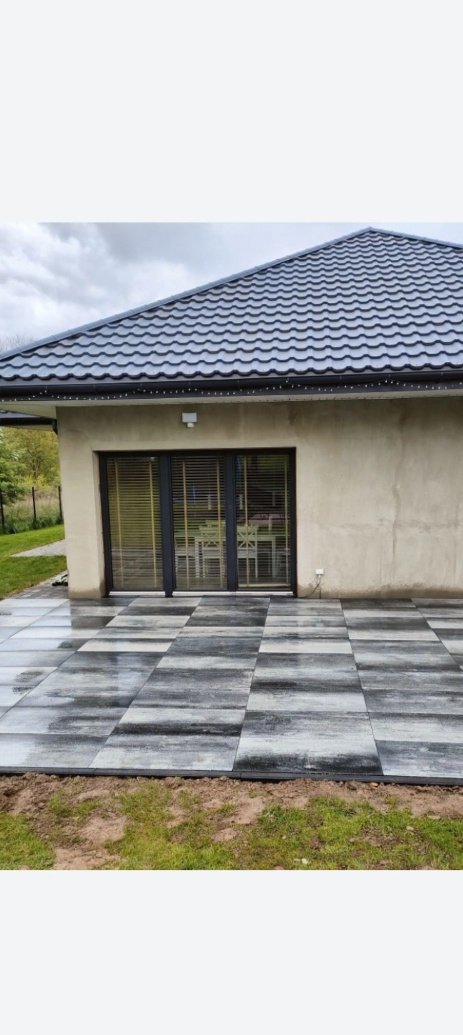 Nowoczesny taras z szarej kostki brukowej ułożonej przy domu z ciemnym dachem. Widoczne duże, prostokątne płyty i staranne wykończenie obrzeży.