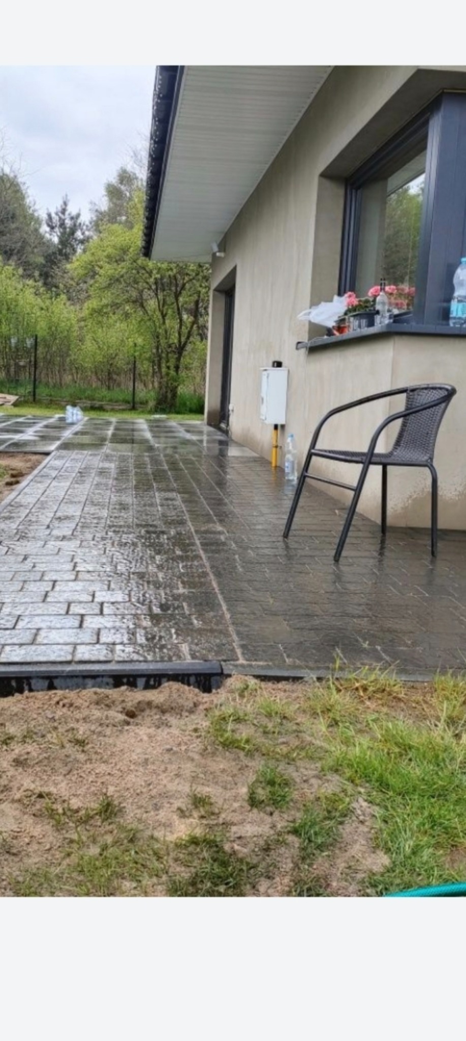 Świeżo ułożona, mokra kostka brukowa prowadząca wzdłuż domu, z widocznym obrzeżem i przygotowanym terenem. Krzesło ogrodowe stoi obok ściany budynku.