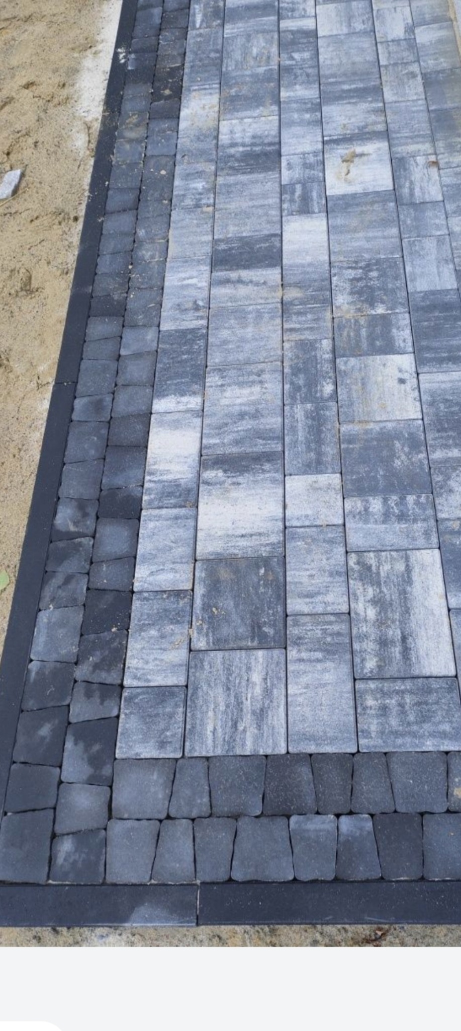 Ułożona kostka brukowa w odcieniach szarości z ciemną obwódką. Widok z góry, ukazujący wzór i fakturę kostki. Częściowo widoczny piasek na obrzeżach.