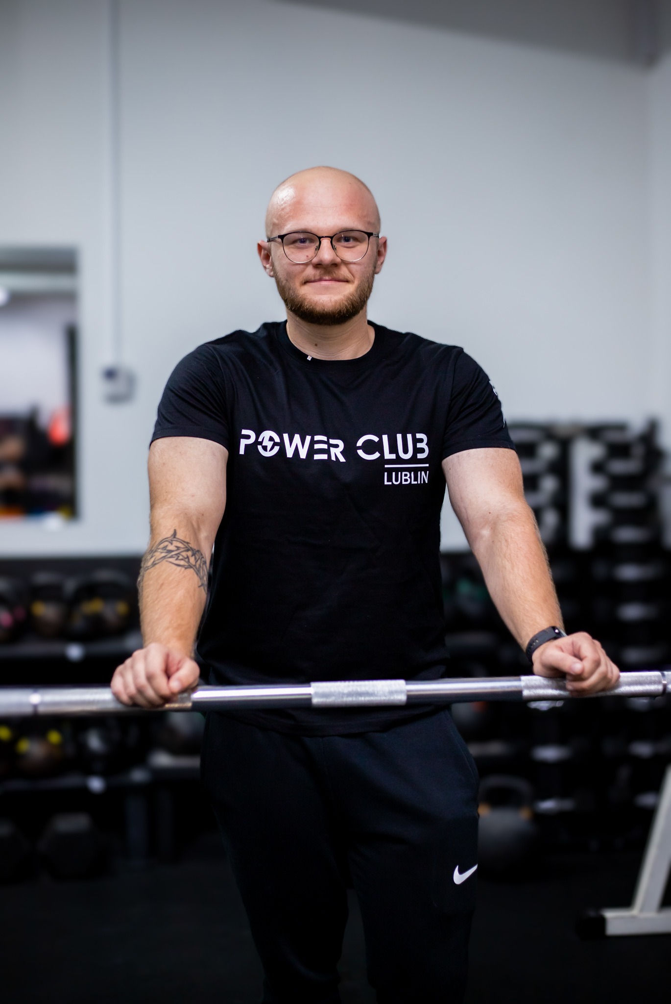 Mężczyzna w czarnej koszulce Power Club Lublin opiera się o sztangę w siłowni, prezentując siłę i kondycję. Widoczne tatuaże na ramieniu.