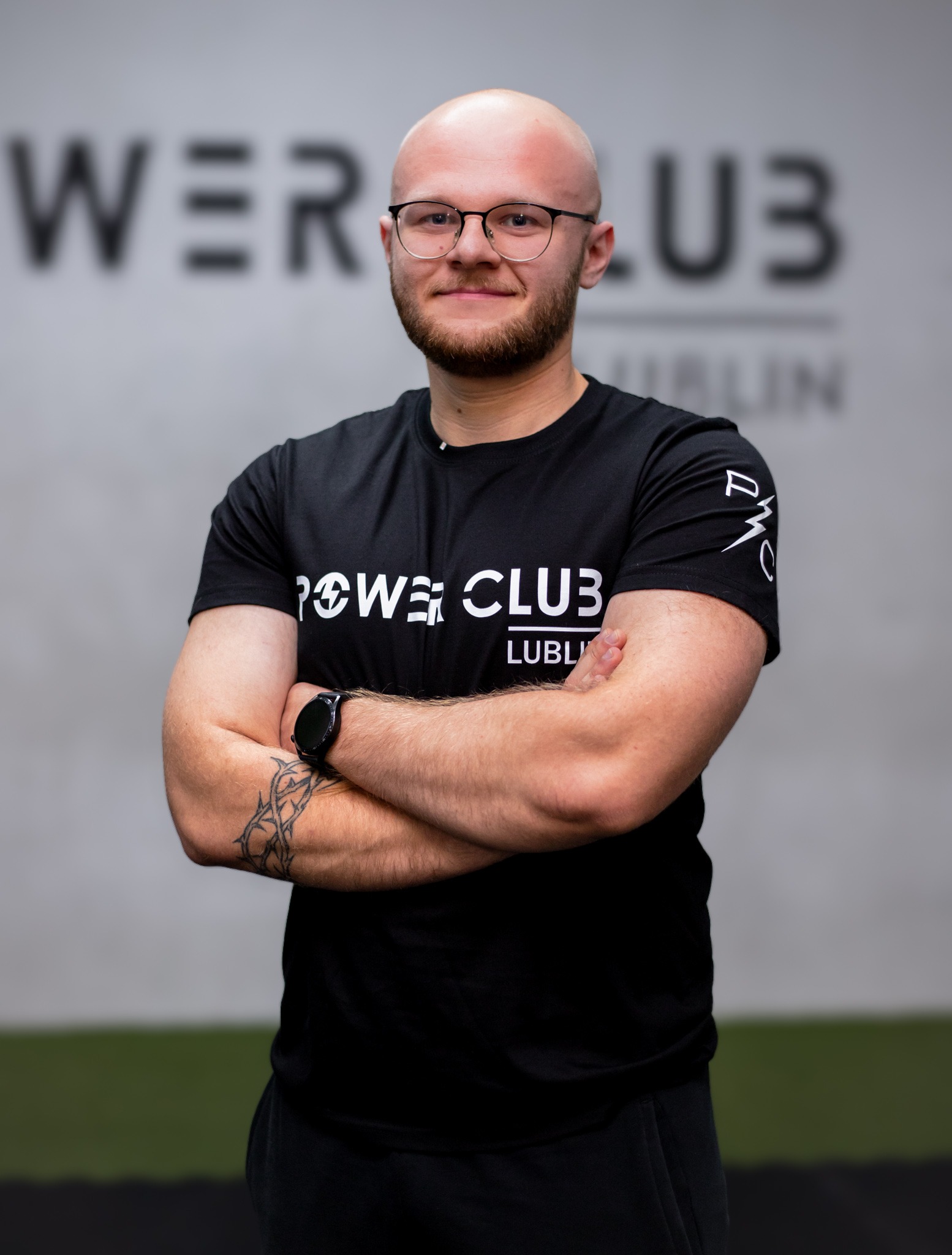 Uśmiechnięty mężczyzna w okularach, z brodą, w czarnej koszulce Power Club Lublin, stoi na tle siłowni z założonymi rękami, prezentując tatuaż na ramieniu.