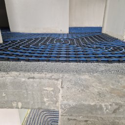 Zeta Profesjonalne Posadzki - Niebieskie rury ogrzewania podłogowego ułożone na czarnej macie z wypustkami, pokryte warstwą szarego kruszywa, widoczne kable elektryczne i surowe ściany.