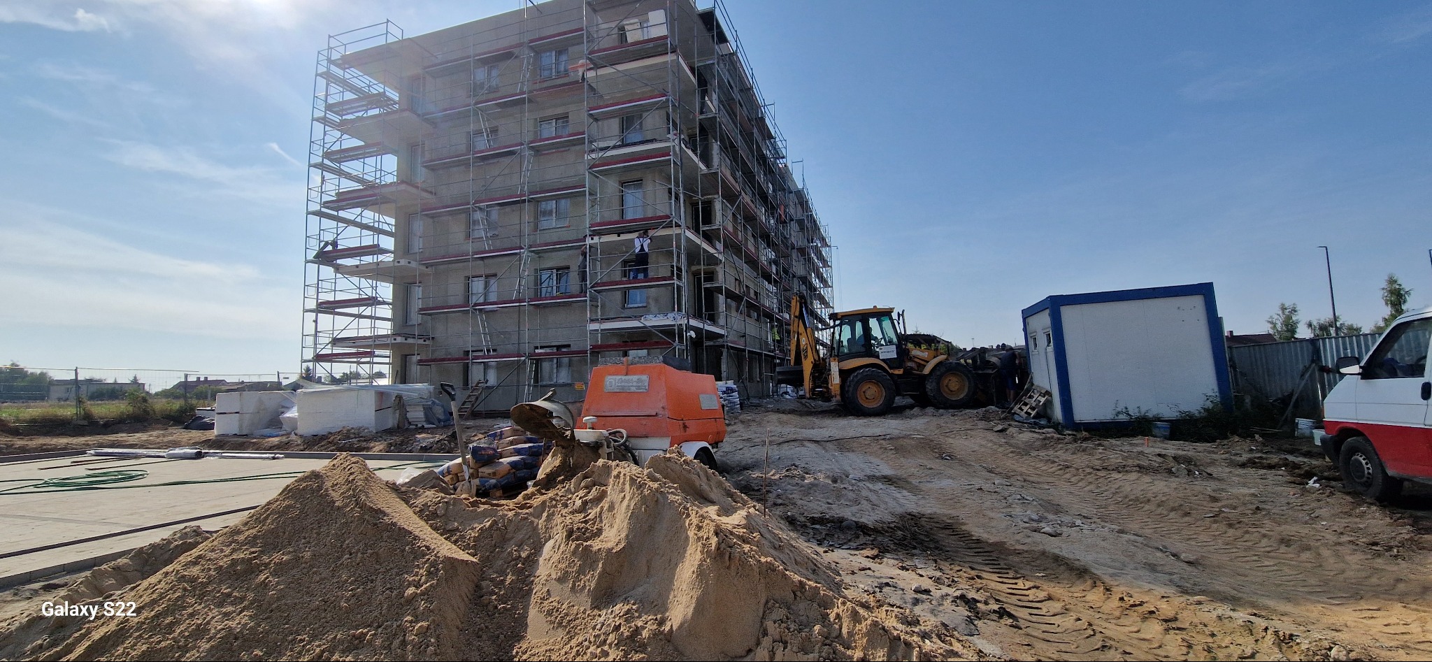 Budowa bloku mieszkalnego w trakcie prac wykończeniowych, widoczne rusztowania, maszyny budowlane i materiały. Słoneczny dzień.