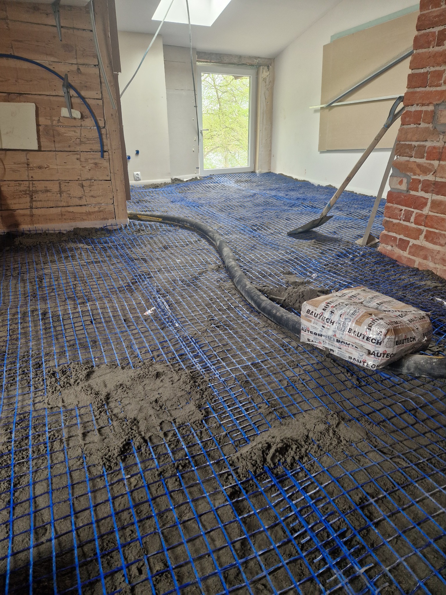 Jastrych półsuchy na siatce z rurami ogrzewania podłogowego w trakcie realizacji w Lipnie. Widoczny worek z zaprawą i szpachelka.