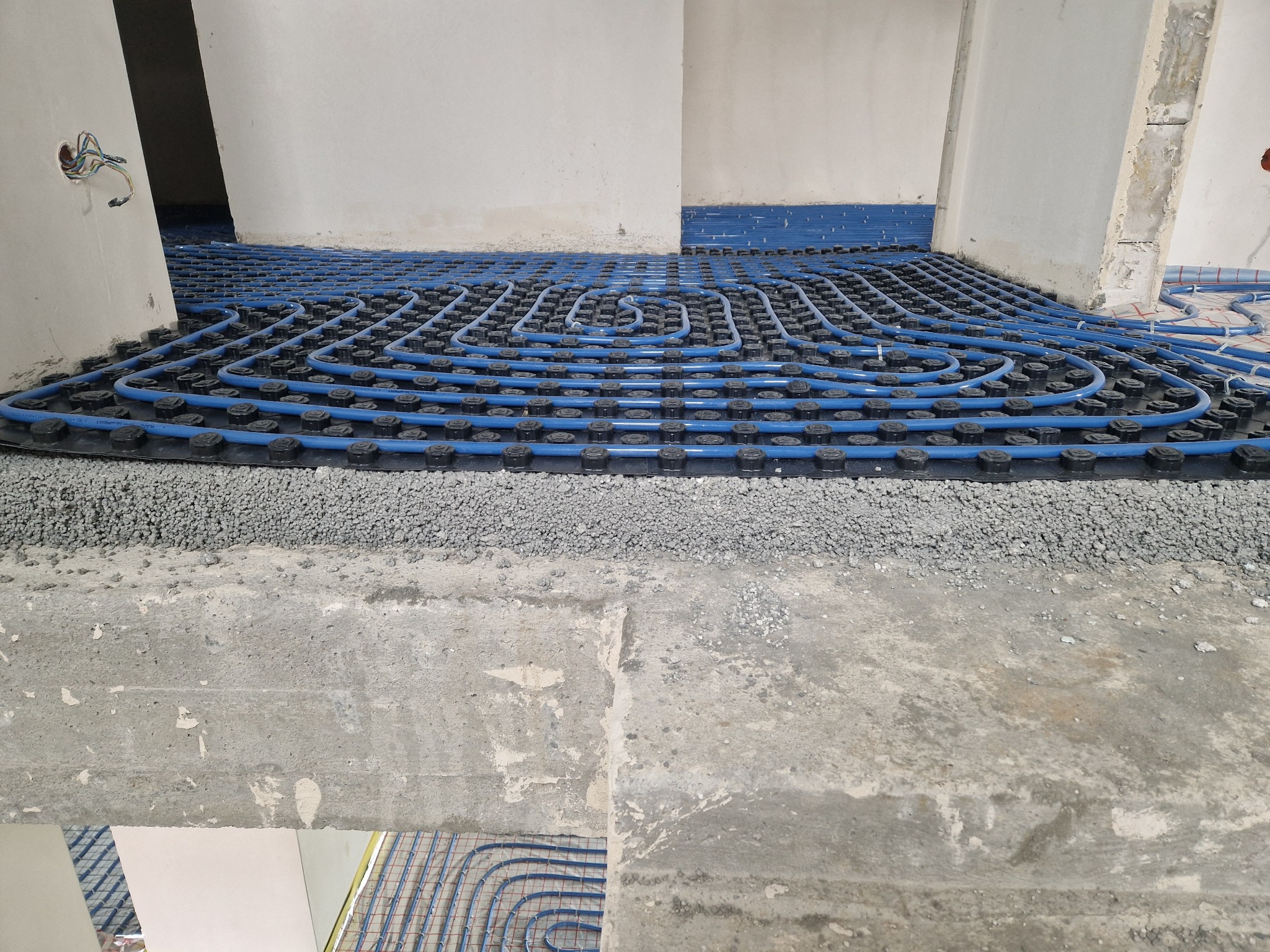 Niebieskie rury ogrzewania podłogowego ułożone na czarnej macie z wypustkami, pokryte warstwą szarego kruszywa, widoczne kable elektryczne i surowe ściany.