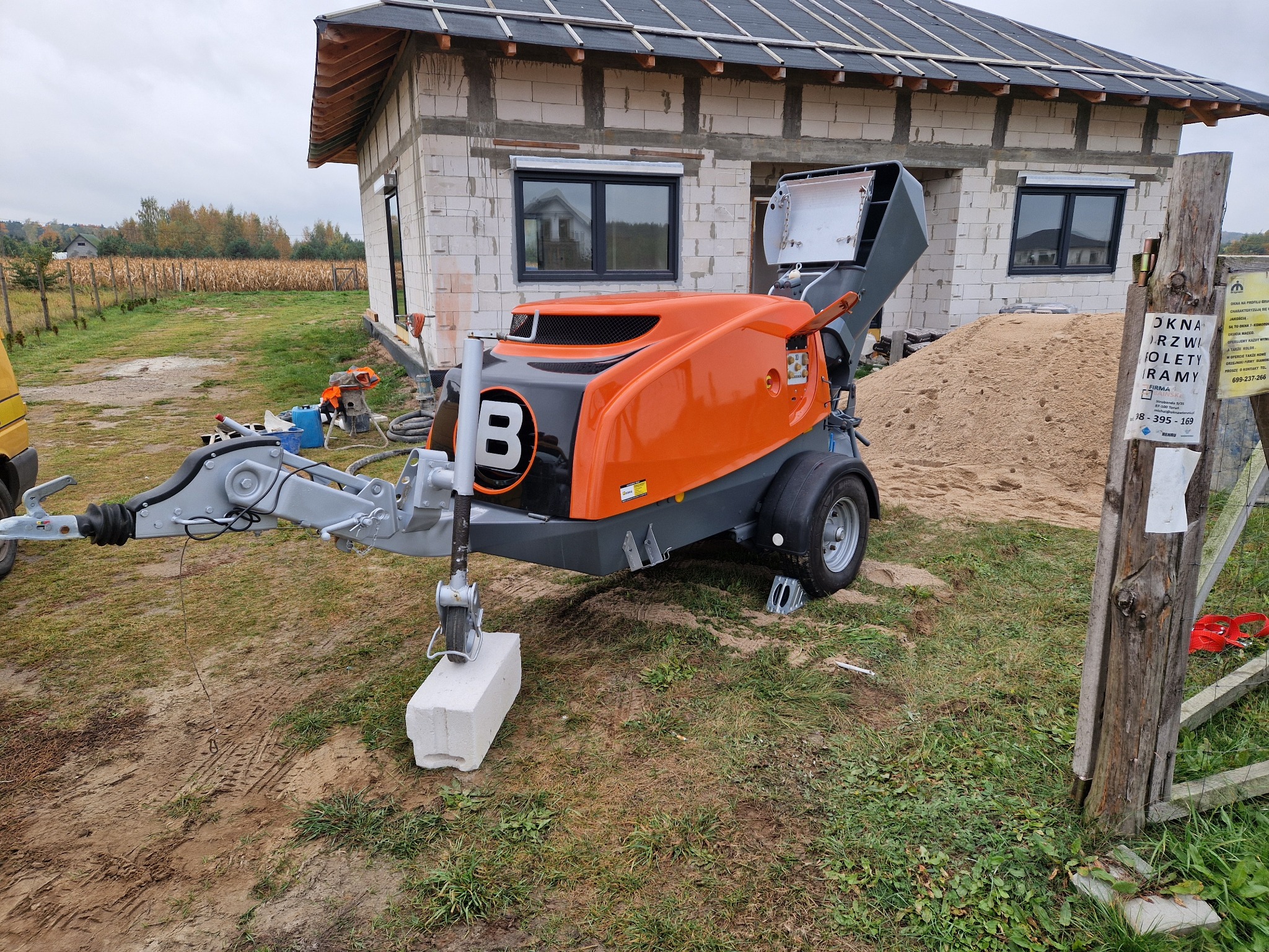 Agregat do posadzek jastrychowych na budowie domu jednorodzinnego w stanie surowym otwartym. Widoczny stos piasku i ogrodzenie z reklamą okien.