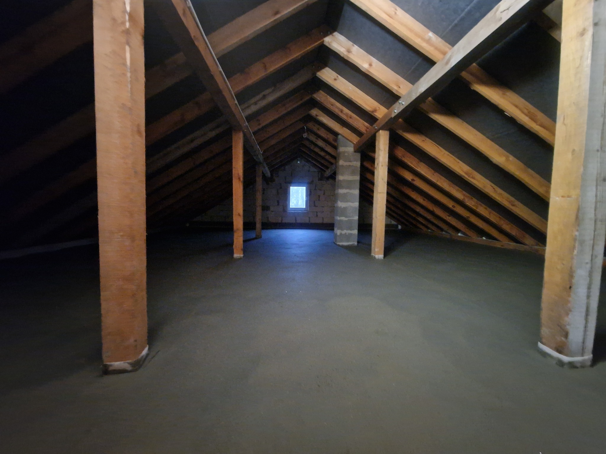 Poddasze z drewnianą konstrukcją i świeżo wylaną, gładką posadzką jastrychową. Widoczne słupy podtrzymujące konstrukcję dachu i okno w oddali.