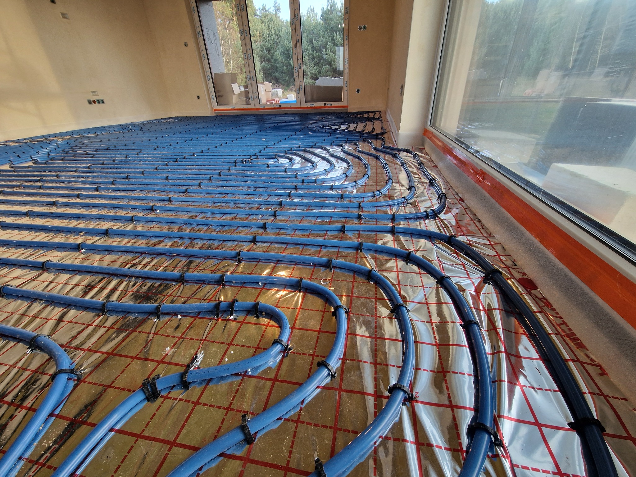 Instalacja ogrzewania podłogowego z niebieskich rur na folii termoizolacyjnej w nowym budynku, widok z góry, duże okna w tle.