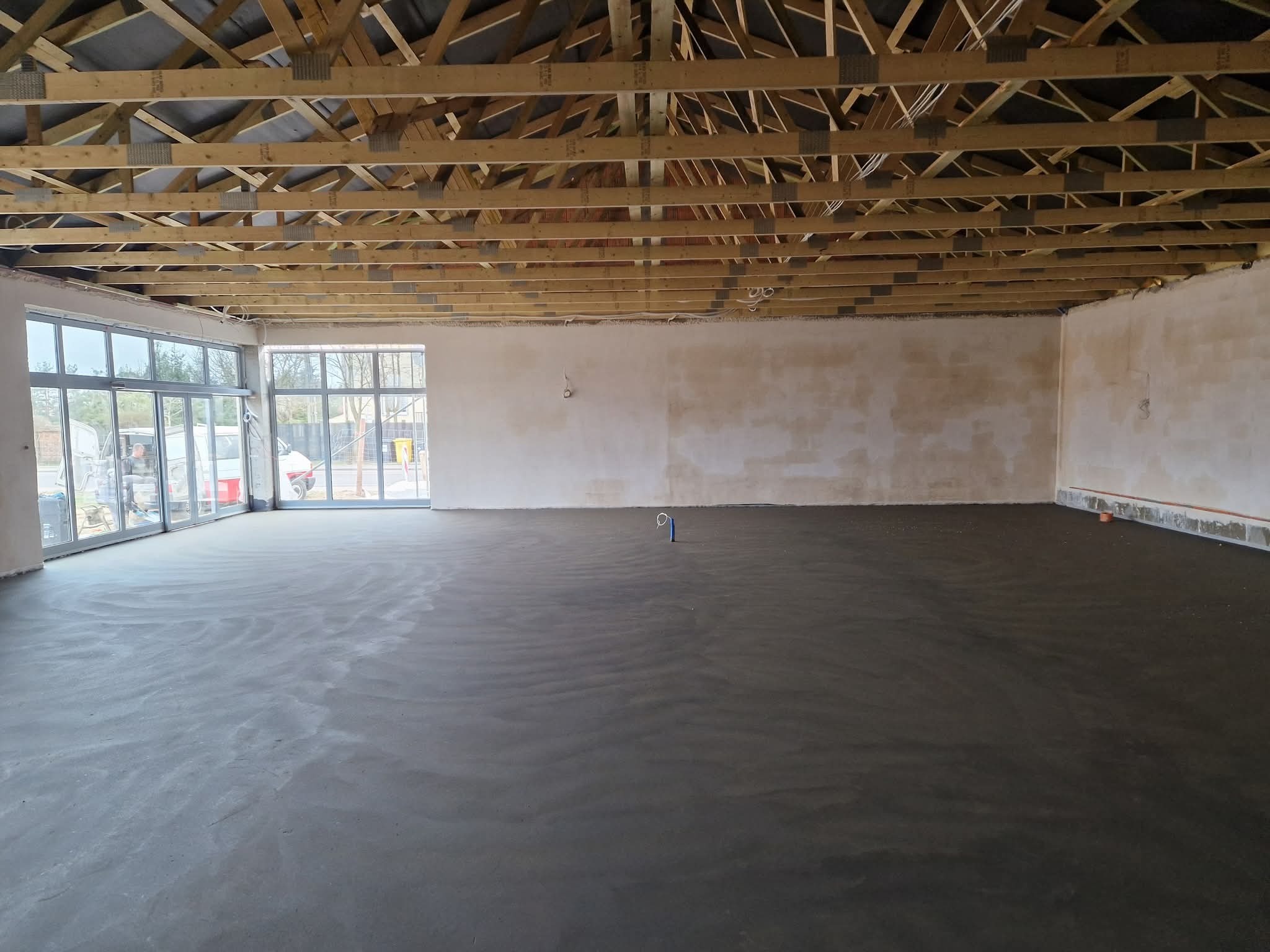 Wnętrze z wylaną, świeżą posadzką jastrychową, widoczne drewniane elementy konstrukcji dachu i duże okna. Szara, gładka powierzchnia podłogi.