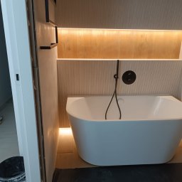 Artbud - Nowoczesna, wolnostojąca wanna z czarną armaturą na tle ściany z podświetlanymi półkami. Widoczny fragment otwartych drzwi i wiadro. Minimalistyczny design łazienki.