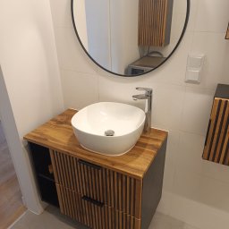 Artbud - Nowoczesna łazienka z drewnianą szafką pod umywalkę i okrągłym lustrem. Umywalka biała, nablatowa. Widoczne płytki na ścianach i podłodze w neutralnych barwach.