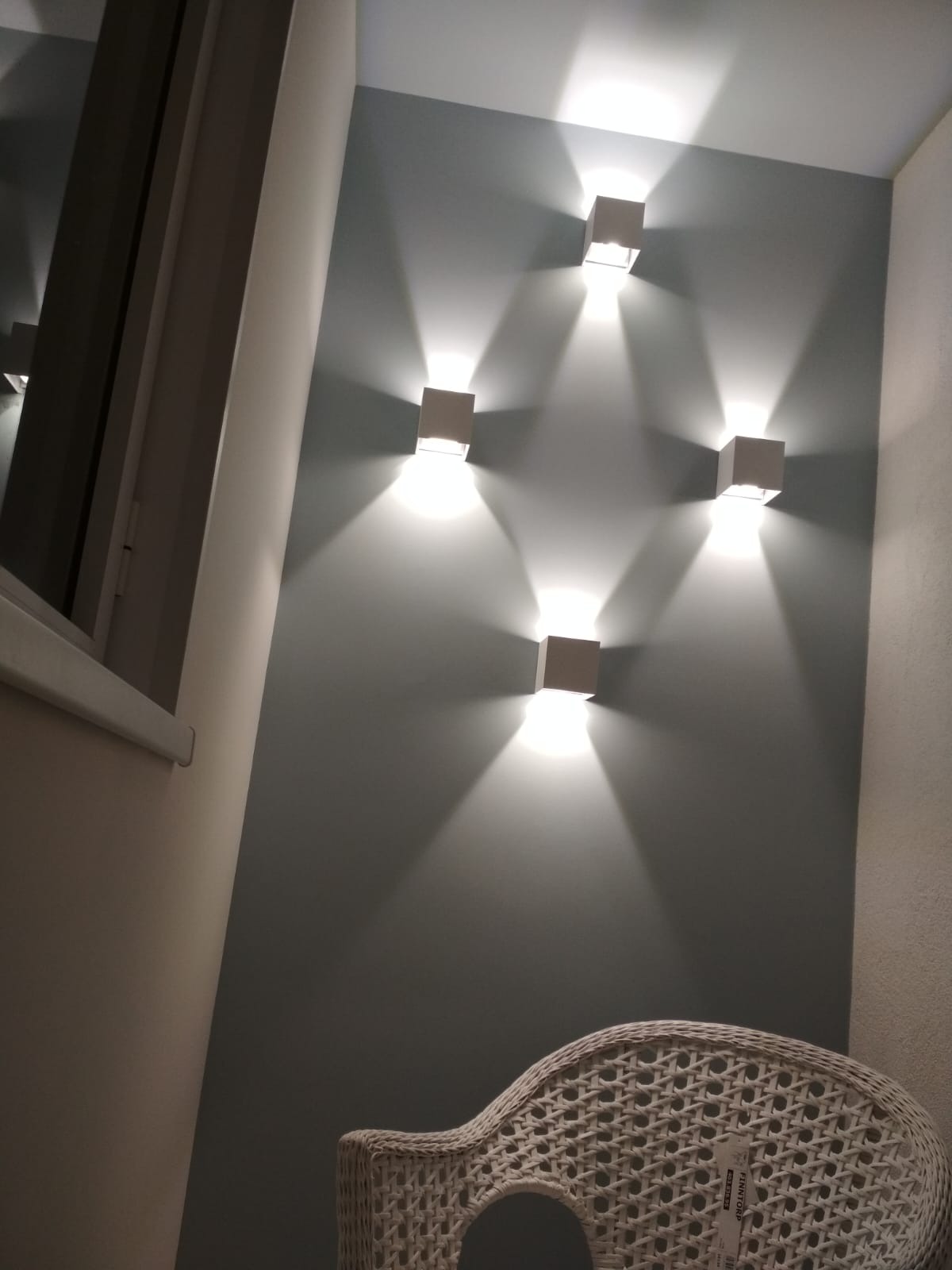 Ściana w odcieniach szarości z czterema kwadratowymi lampami ściennymi, rzucającymi jasne światło. W rogu widoczny fragment białego, wiklinowego krzesła.